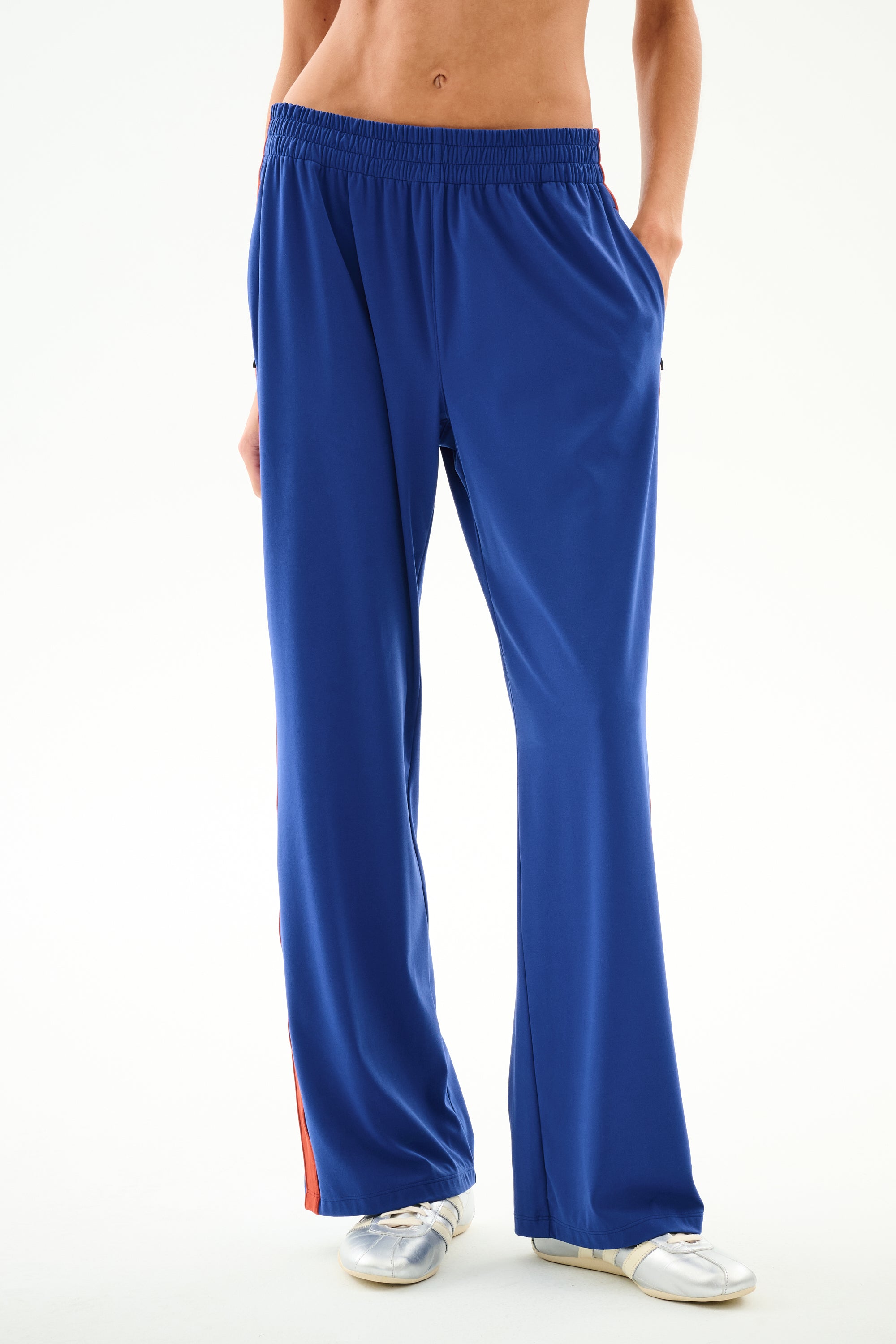 Model wears SPLITS59 Maxie Rigor Track Pant - Cobalt/Terracotta, with red side stripes, white sneakers, hands in pockets; shown waist down on white background.