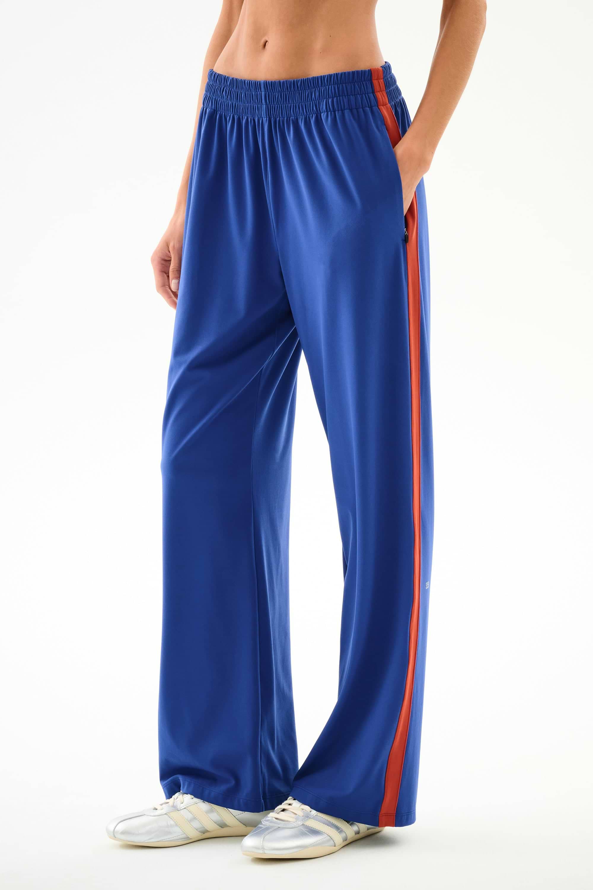Model wears SPLITS59 Maxie Rigor Track Pant - Cobalt/Terracotta with a red stripe and white sneakers, hands in pockets, shown waist down against a plain background.