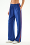 Model wears SPLITS59 Maxie Rigor Track Pant - Cobalt/Terracotta with a red stripe and white sneakers, hands in pockets, shown waist down against a plain background.