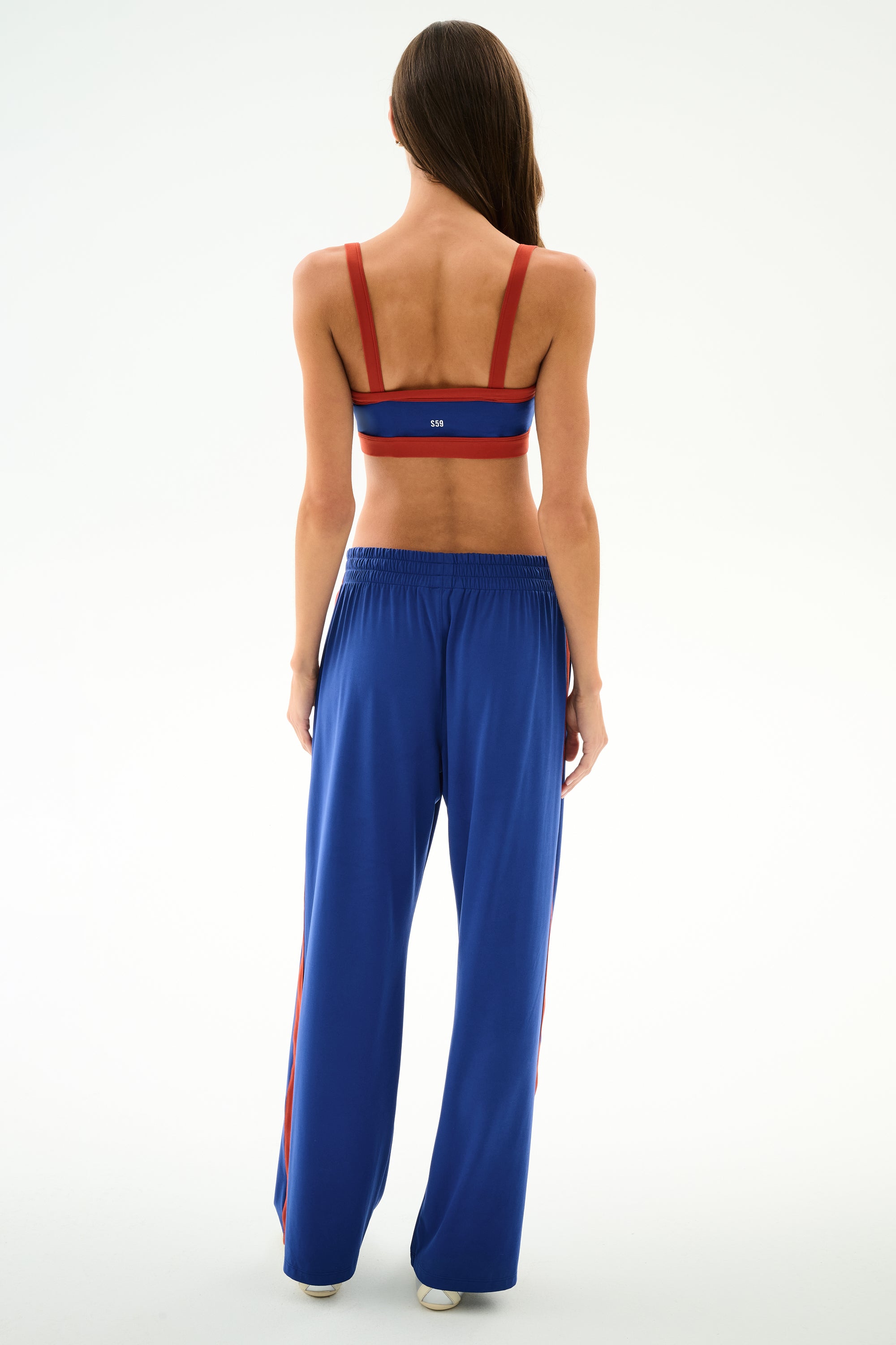 A woman faces away, wearing a red sports bra and SPLITS59 Maxie Rigor Track Pant - Cobalt/Terracotta, against a plain white background.