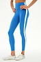A person stands against a plain background, hands on hips, wearing SPLITS59’s Clare High Waist Rigor 7/8 leggings in Classic Blue/White—crafted from a soft nylon spandex blend and featuring a distinctive white side stripe—paired with white sneakers.