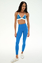A woman wearing the SPLITS59 Sage Rigor Bra in Classic Blue/White stands against a plain white background, paired with matching blue leggings and white sneakers. The outfit is crafted from stretchy nylon spandex fabric for comfort and flexibility.