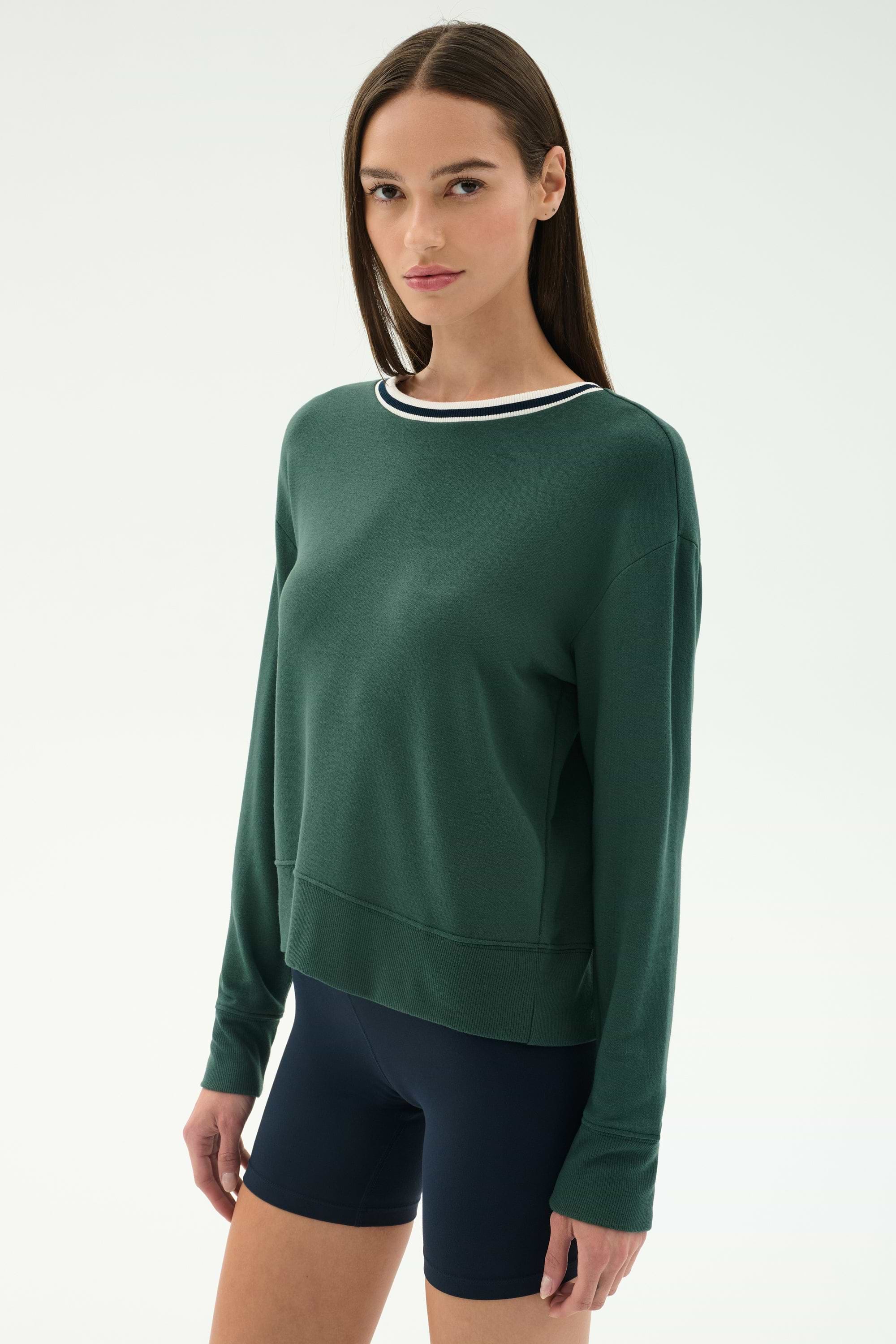 A woman with straight brown hair wears the SPLITS59 Sonja Fleece Sweatshirt W/ Stripe in Military and navy spandex bicycle shorts, standing against a plain light background.