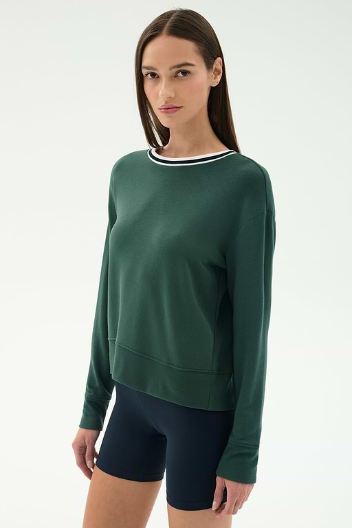 A woman with straight brown hair wears the SPLITS59 Sonja Fleece Sweatshirt W/ Stripe in Military and navy spandex bicycle shorts, standing against a plain light background.