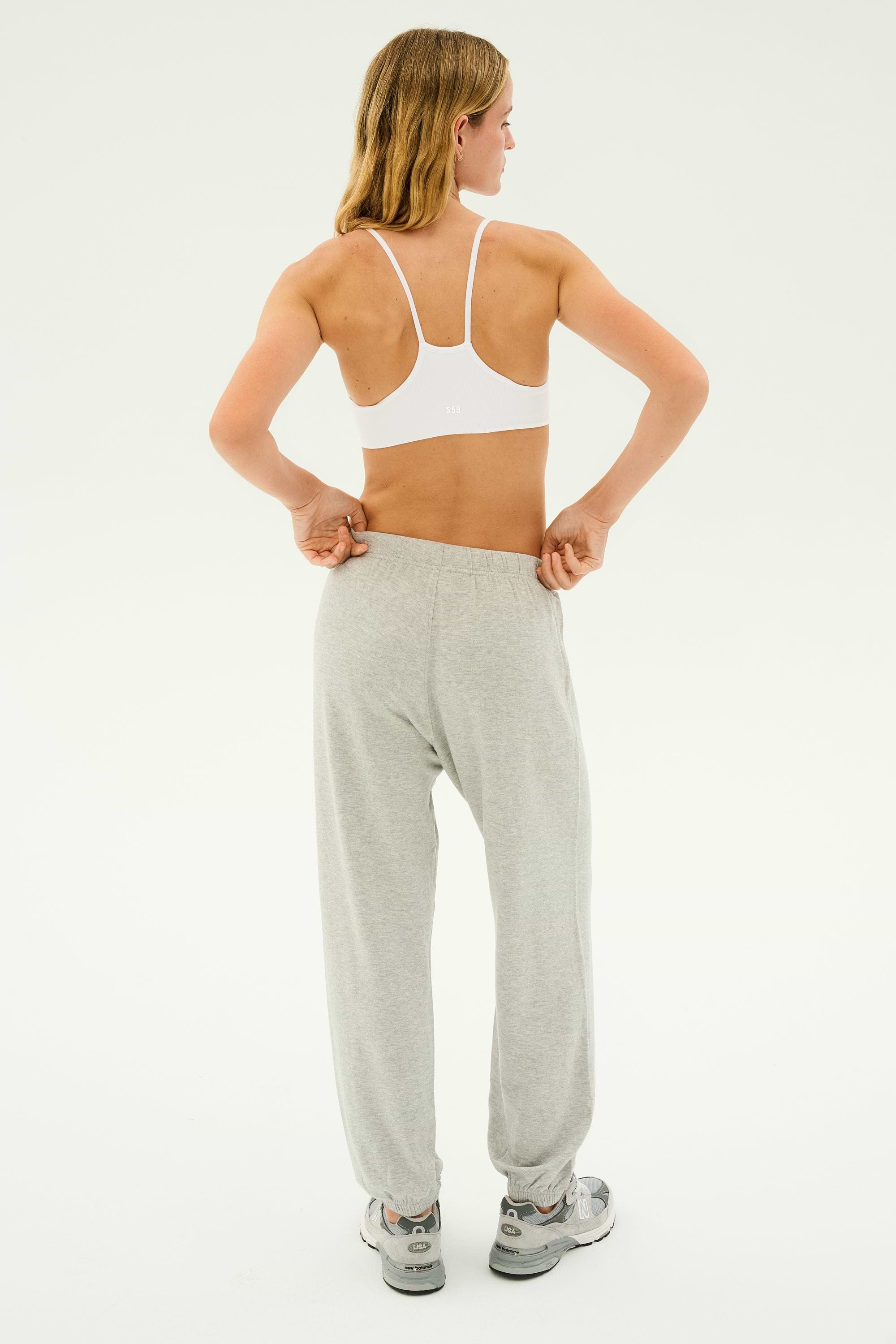 A person in a white sports bra is standing and adjusting the waistband of their Andie Oversized Fleece Sweatpant in Heather Grey by SPLITS59. The person is facing away from the camera with their head turned slightly to the side, making it perfect for cool-weather workouts.
