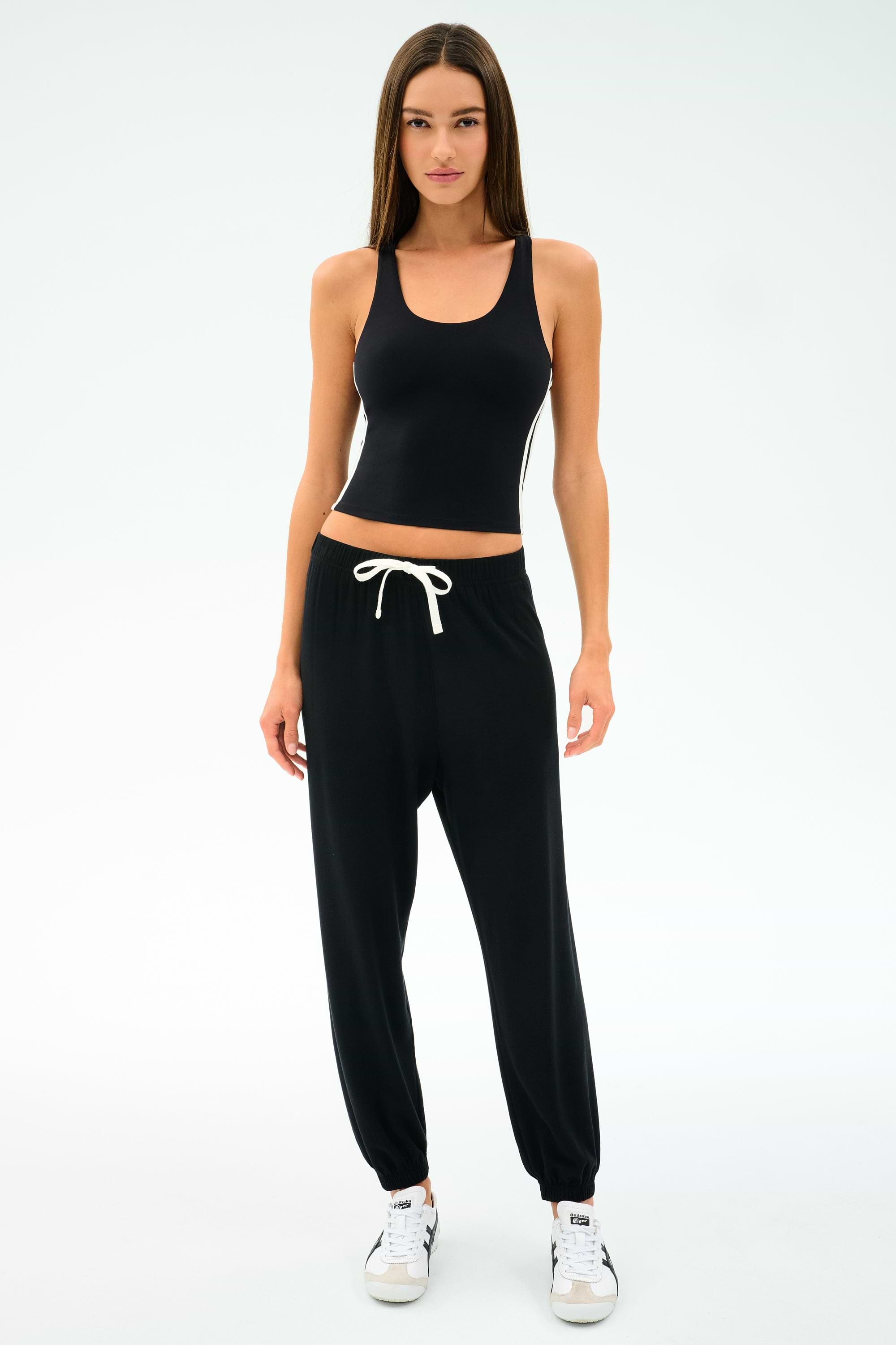 A woman faces forward in the SPLITS59 Ella Airweight Tank - Black/White, paired with black jogger pants and white sneakers, set against a plain white background.