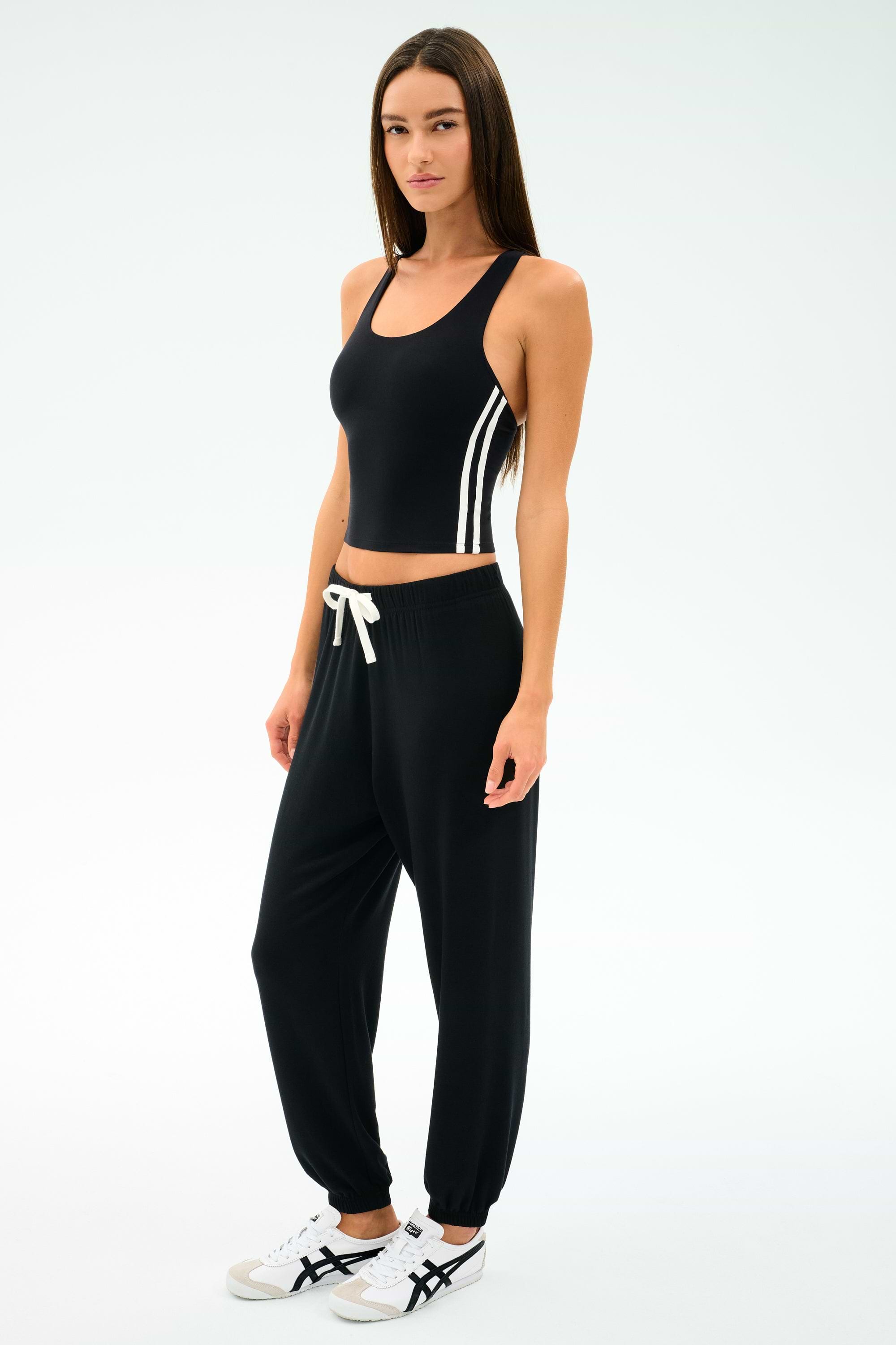 A woman faces forward on a white background, wearing the SPLITS59 Ella Airweight Tank in Black/White with side stripes, black nylon-spandex jogger pants, and white sneakers.