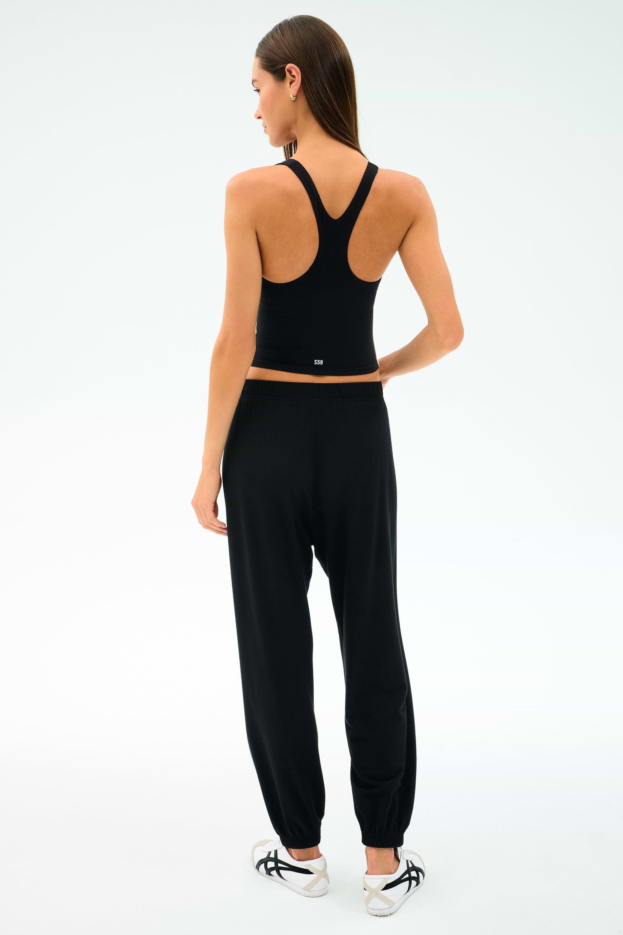 A woman faces away wearing the SPLITS59 Ella Airweight Tank in Black/White and jogger pants made from a soft nylon-spandex blend, paired with white sneakers featuring black accents.