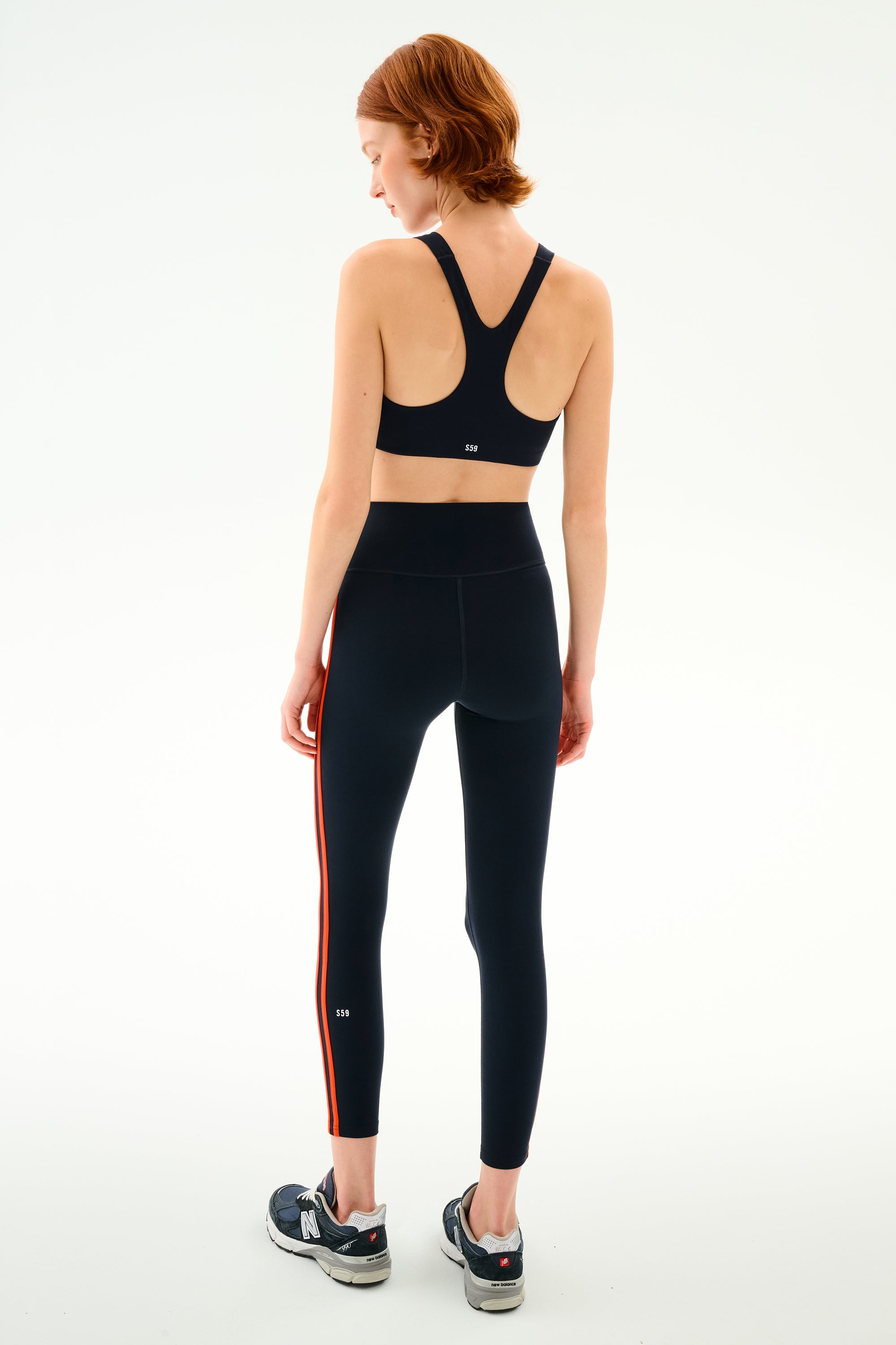 A person seen from behind wearing a black nylon racerback sports bra and SPLITS59 Ella High Waist Airweight 7/8 - Indigo/Valencia leggings with orange side stripes, paired with dark athletic sneakers.