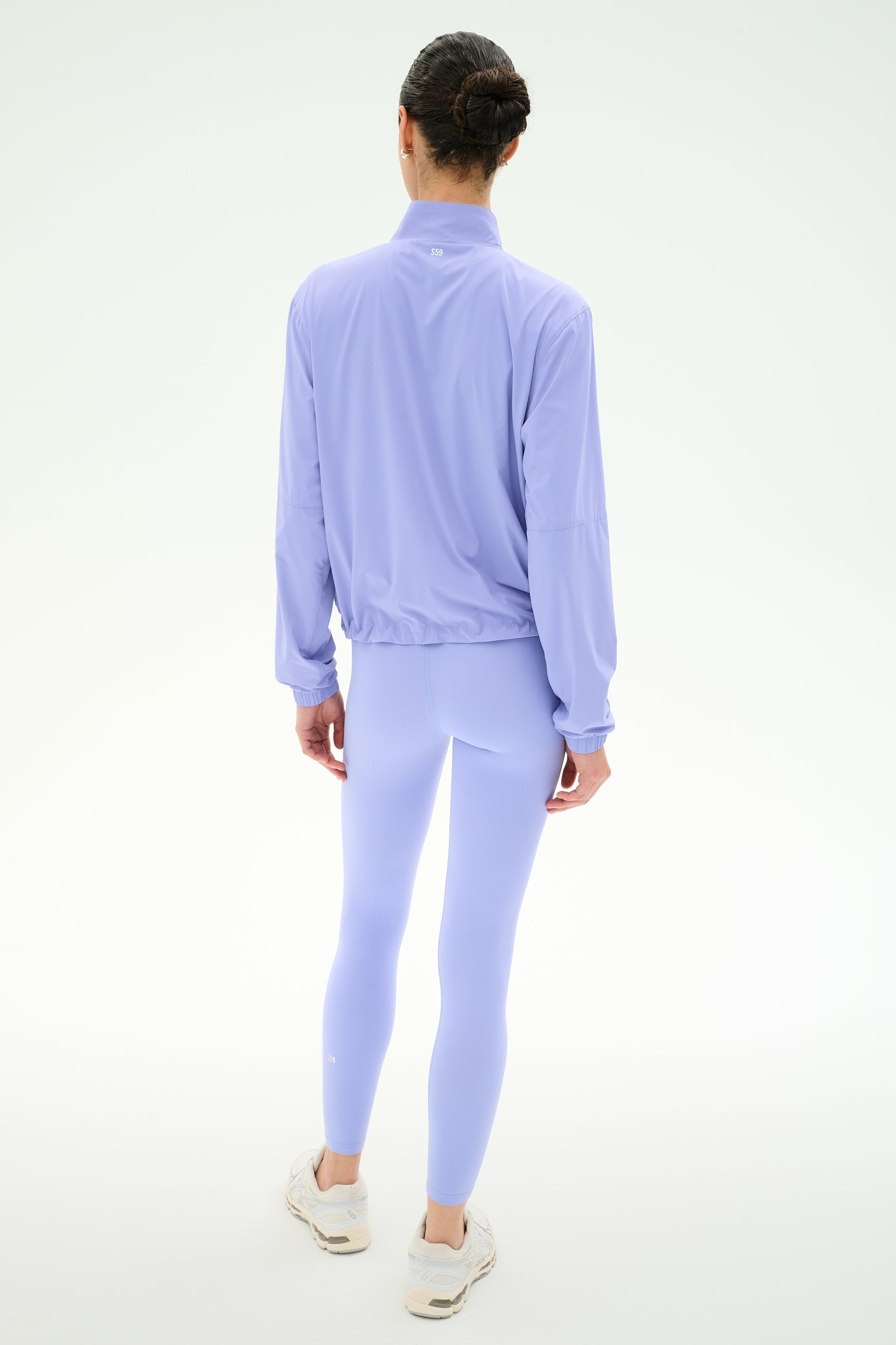 A person stands with their back to the camera, wearing the SPLITS59 Lyle Woven Nylon Pullover in Purple Haze/Creme and matching leggings with white sneakers against a plain white background.