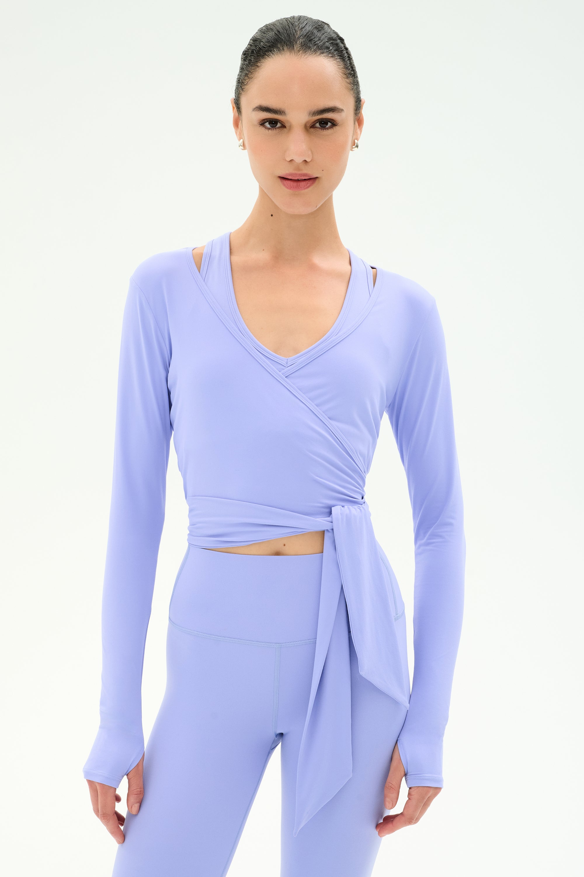A woman wearing the SPLITS59 Bella Airweight Lite Wrap top in Purple Haze with matching high-waisted leggings stands against a plain white background.