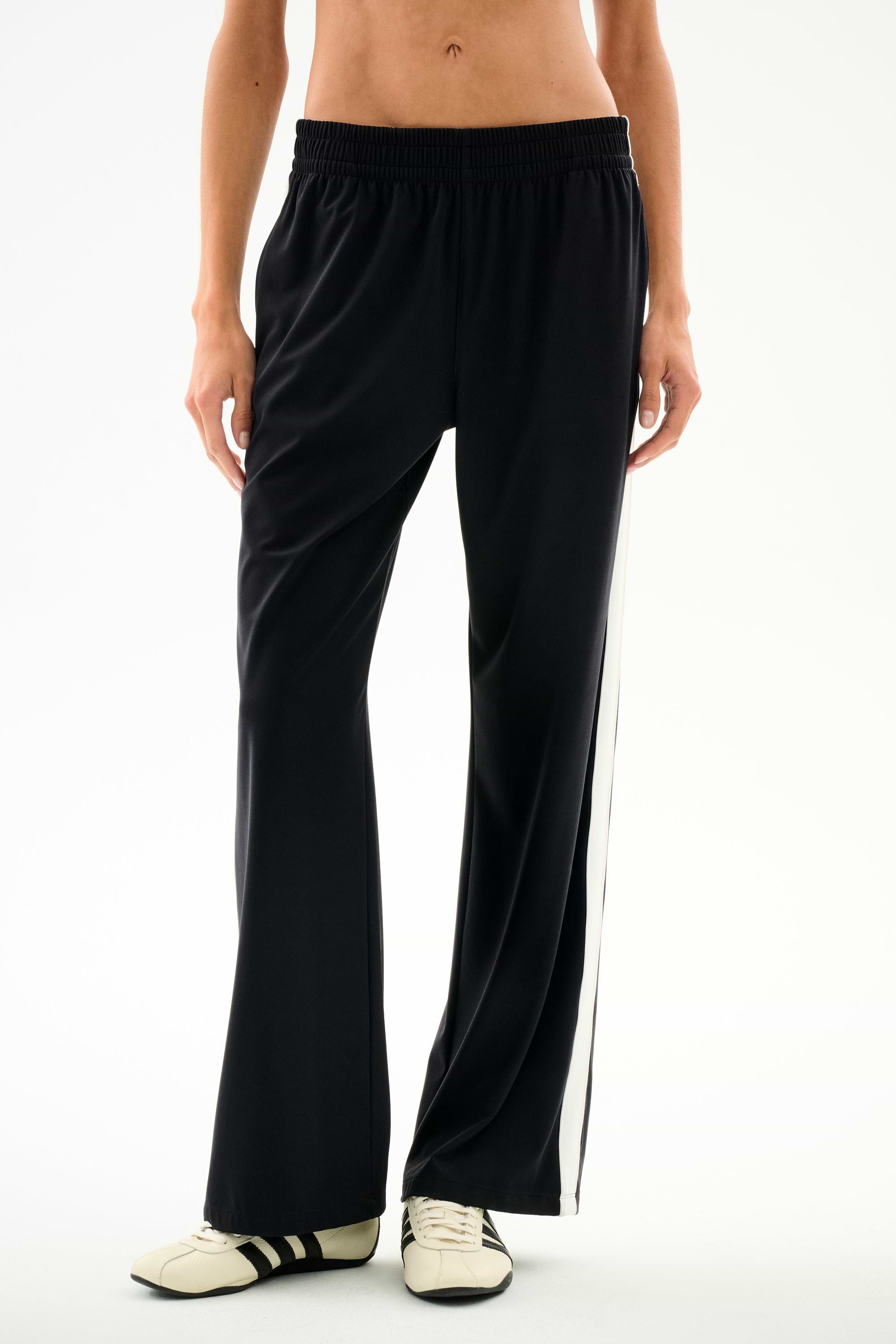 Black wide-leg Maxie Rigor Track Pant - Black/White with a white side stripe, worn with black and white sneakers.