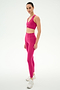 A person stands sideways wearing the SPLITS59 Airweight Bra with Ruching in Sangria and matching leggings with white athletic shoes, set against a plain white background.
