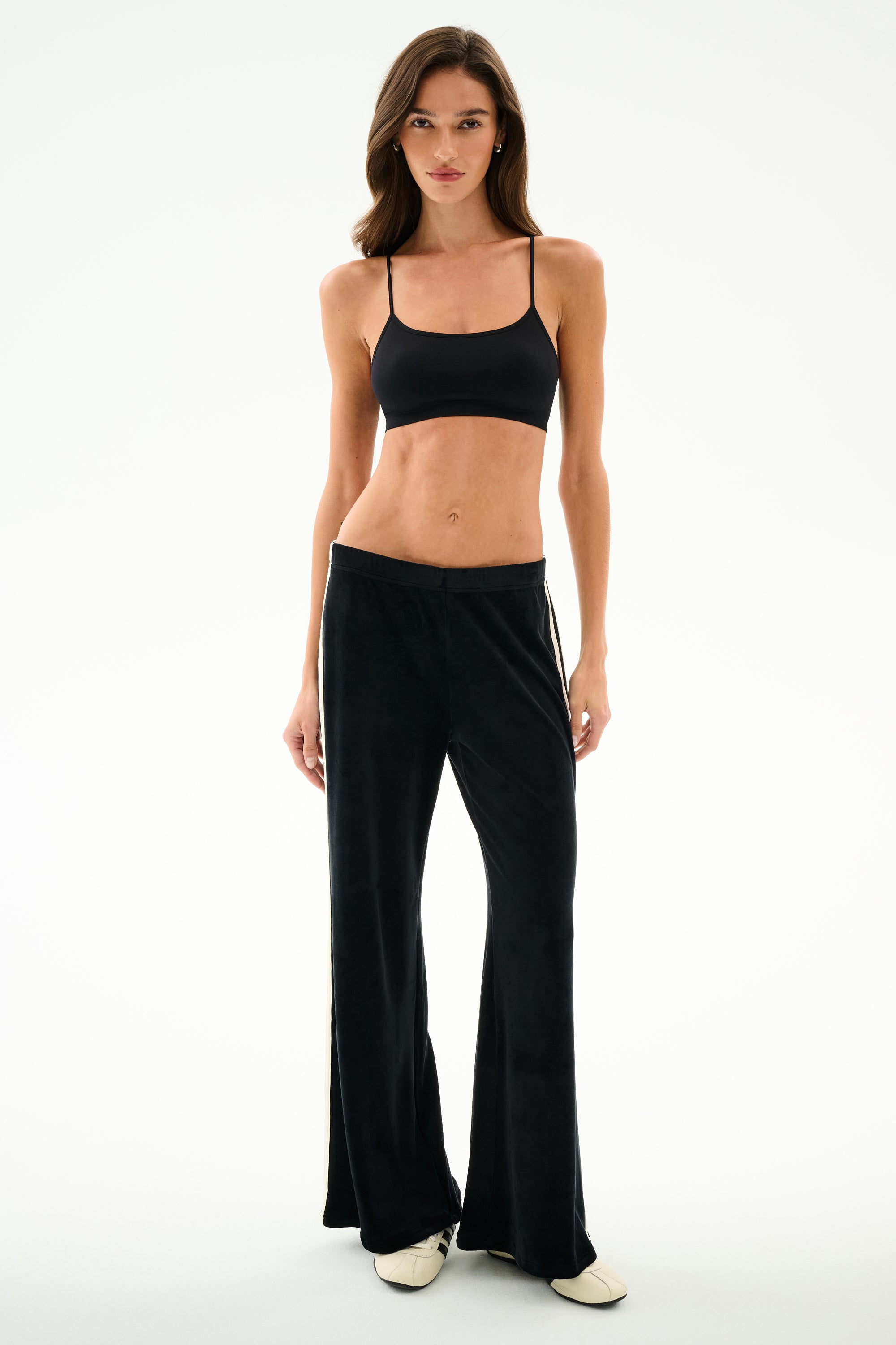A woman stands against a white background wearing SPLITS59 Heritage Velour Pant - Black/Creme, a black crop top, and white shoes.