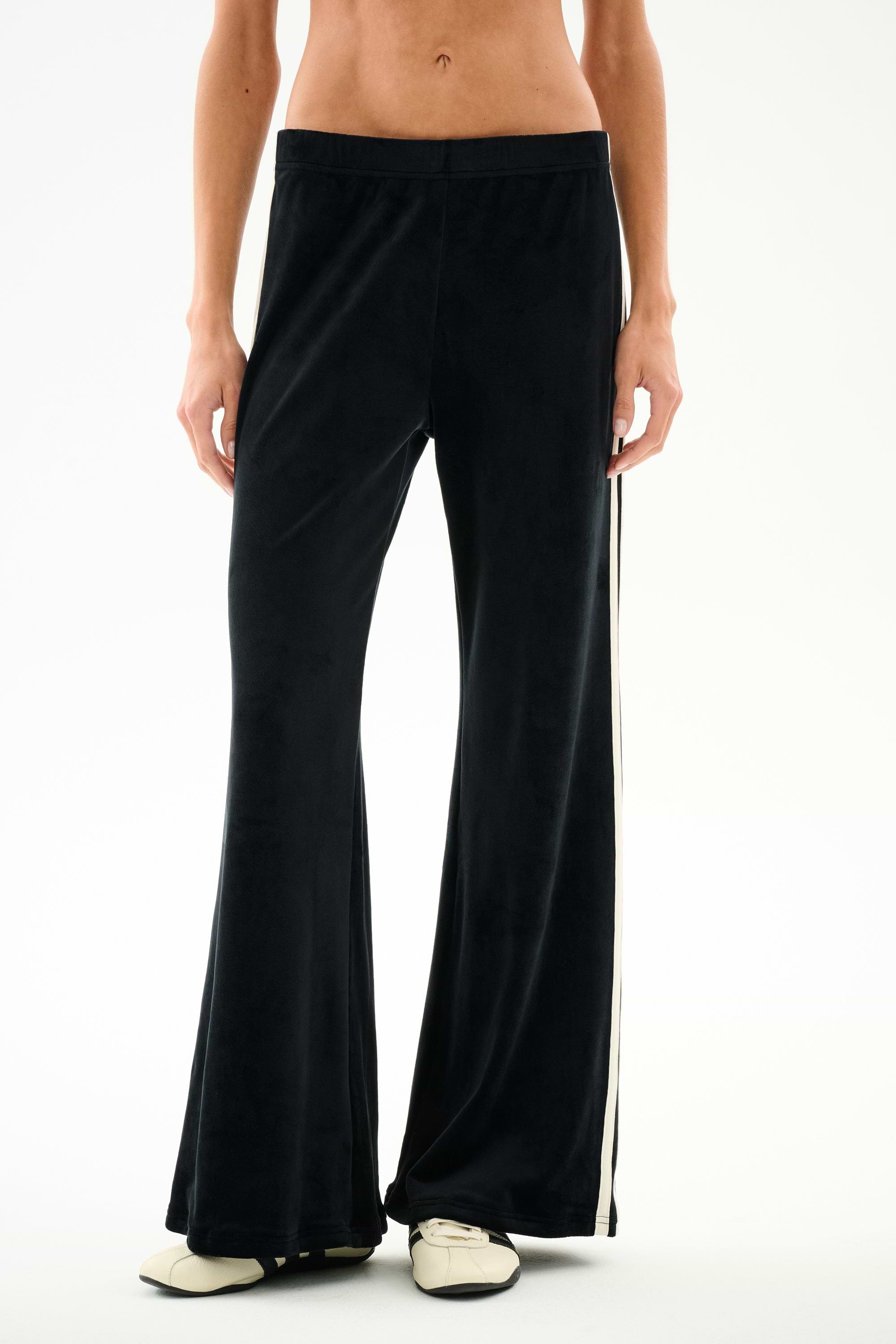 Model showcases SPLITS59 Heritage Velour Pant - Black/Creme, featuring a white side stripe and flared silhouette; image focuses on lower torso and legs against a white background.