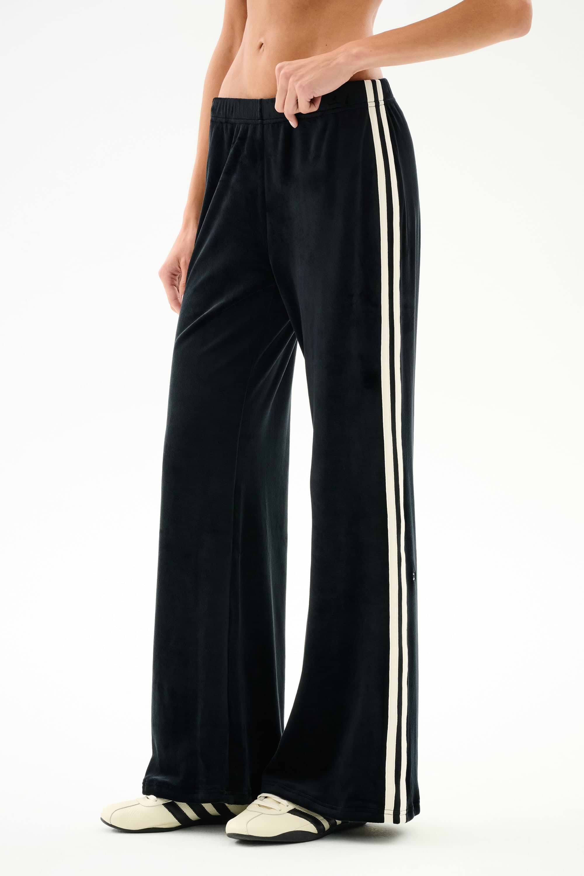 Person wearing SPLITS59 Heritage Velour Pant - Black/Creme with white stripes and cream sneakers, standing against a plain white background.