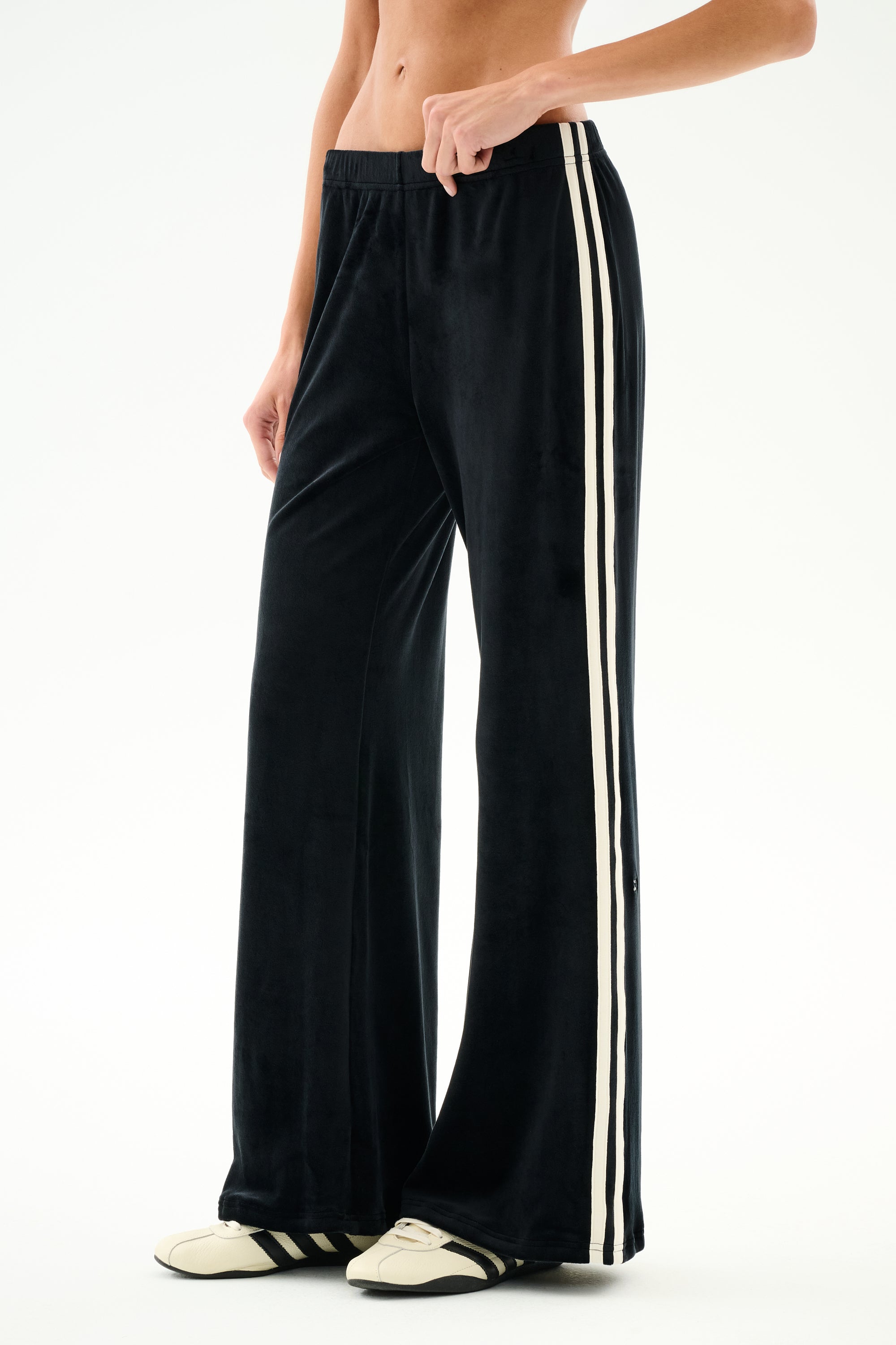 Person wearing SPLITS59 Heritage Velour Pant - Black/Creme with white stripes and cream sneakers, standing against a plain white background.