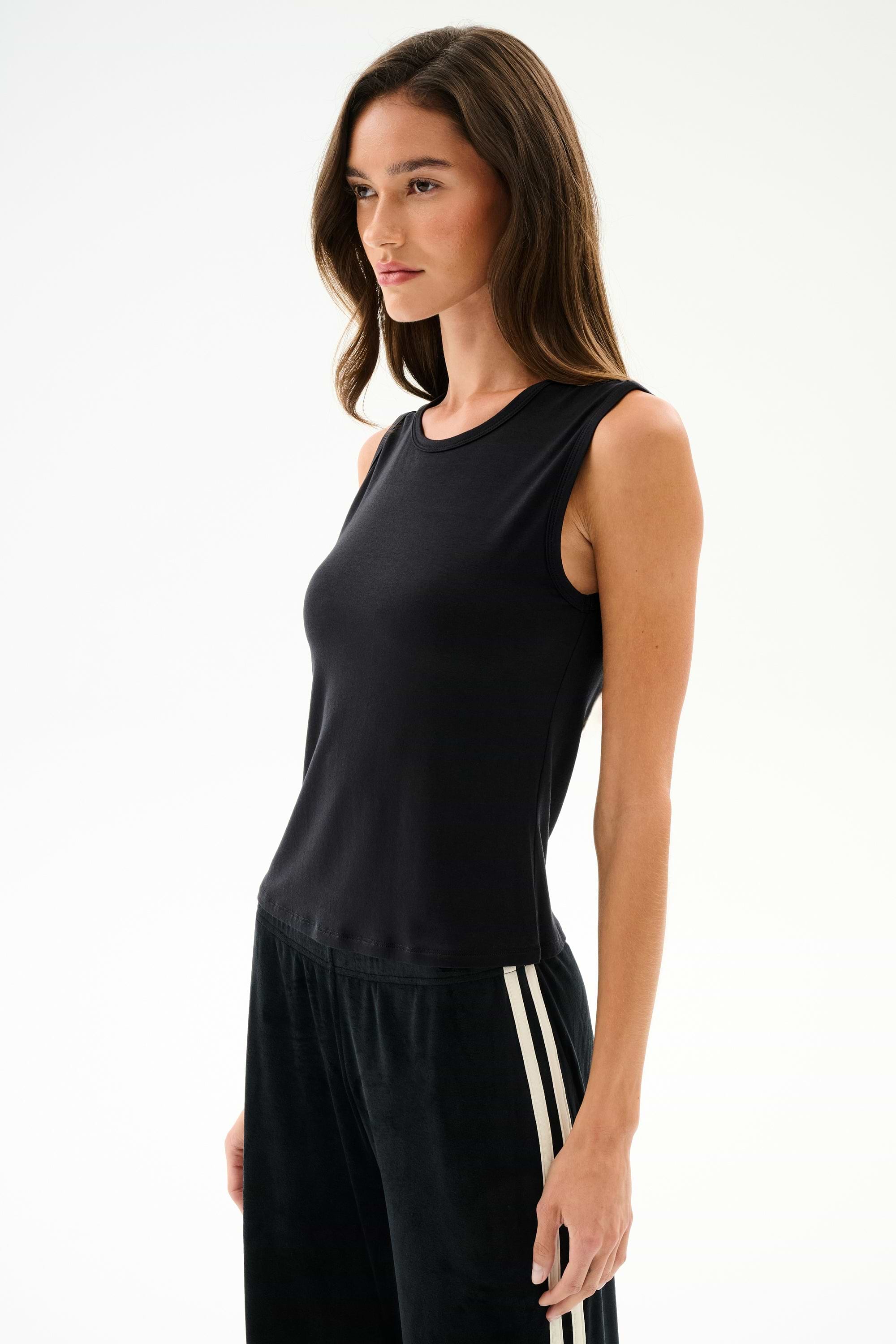 A woman wears the SPLITS59 Nikki Jersey Tank - Black with black pants featuring white stripes, standing against a plain light background, facing forward neutrally.