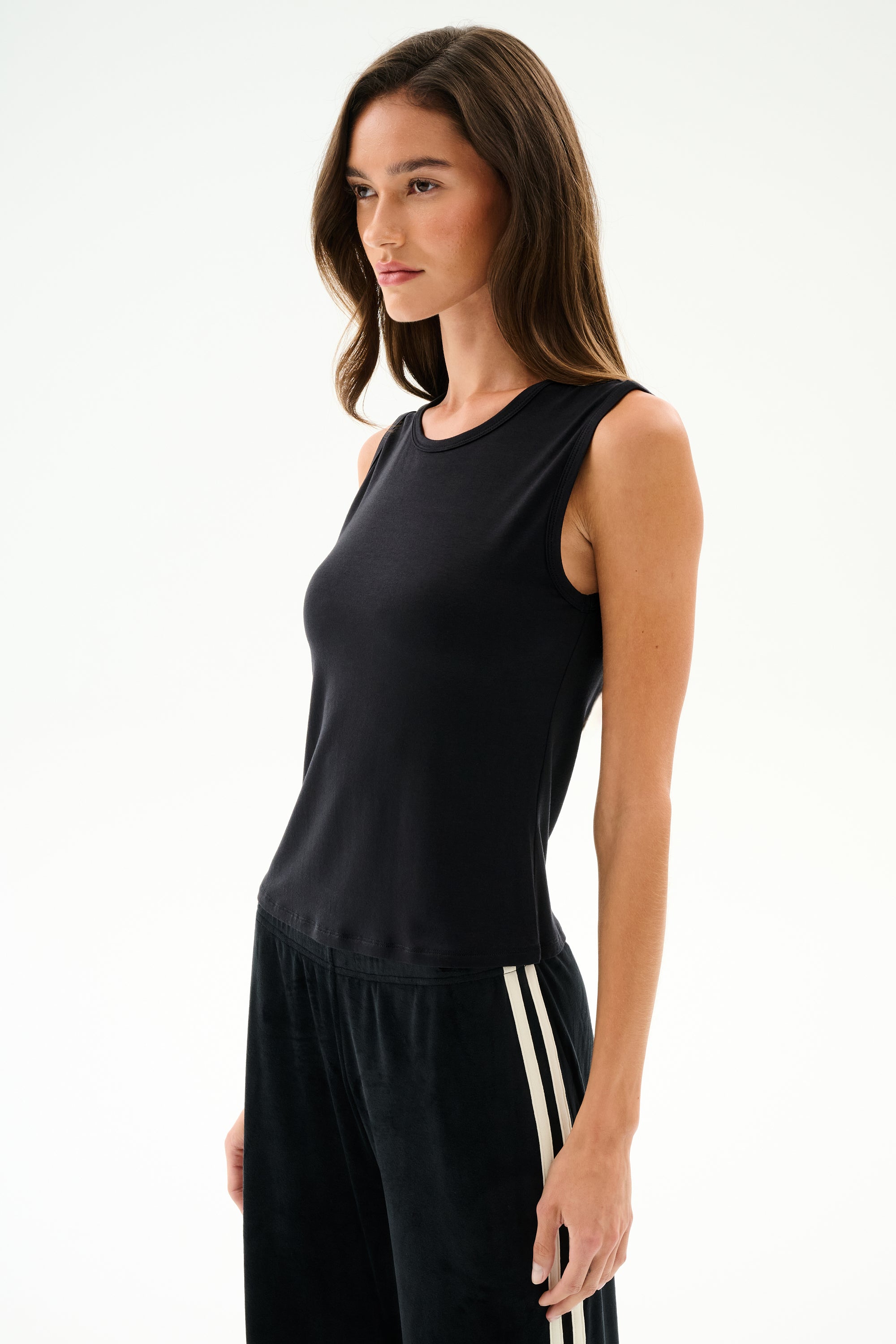 A woman wears the SPLITS59 Nikki Jersey Tank - Black with black pants featuring white stripes, standing against a plain light background, facing forward neutrally.