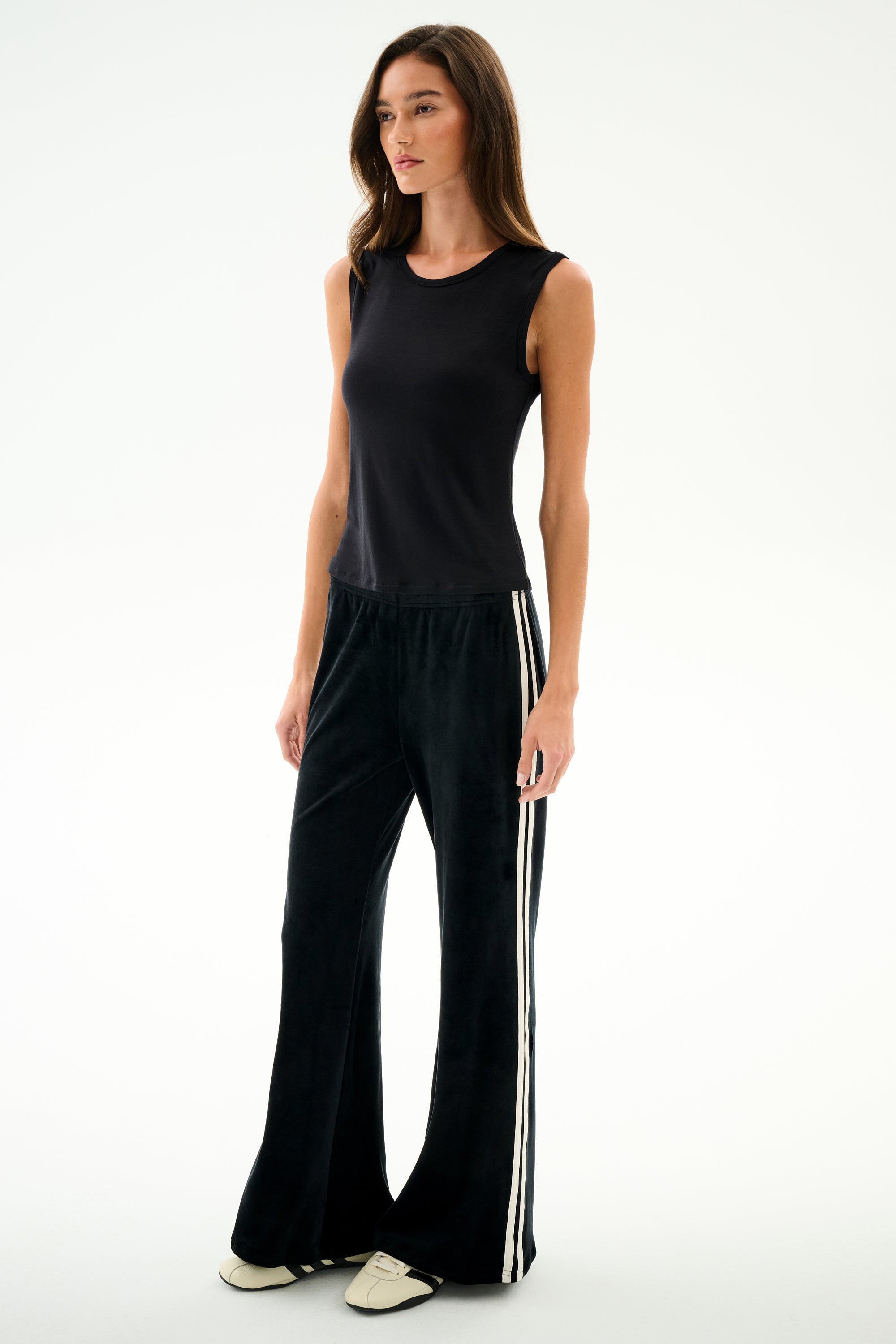 A woman wears the SPLITS59 Nikki Jersey Tank - Black with black flared viscose pants featuring white side stripes and white shoes, standing against a plain white background.