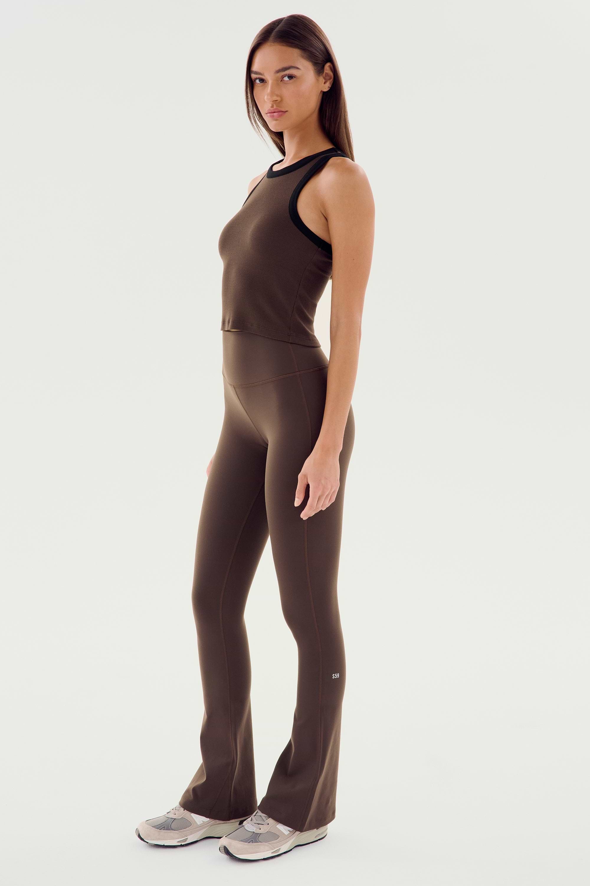 A woman stands in a side pose wearing the SPLITS59 Raquel High Waist Flared Legging in Dark Chocolate with a matching athletic top and light-colored sneakers against a plain white background.