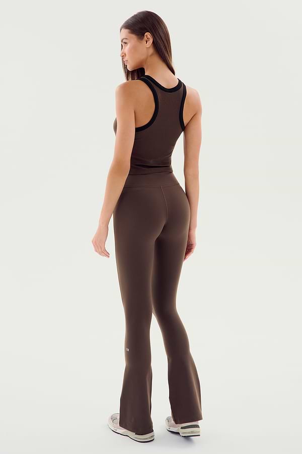 A woman faces away, wearing the SPLITS59 Raquel High Waist Flared Legging in Dark Chocolate, a sleeveless brown top, and light sneakers, posed against a plain white background.