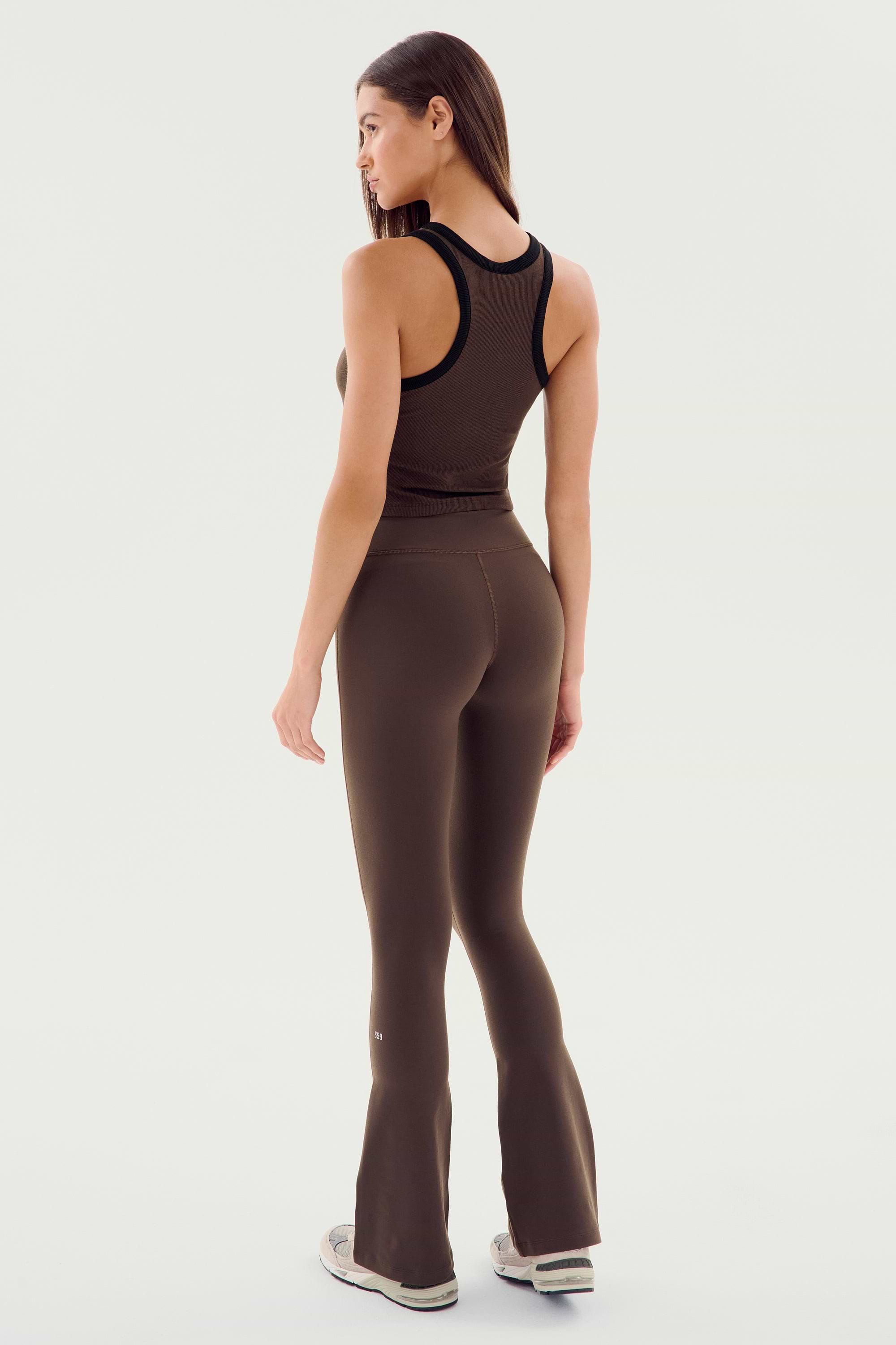 A woman faces away, wearing the SPLITS59 Raquel High Waist Flared Legging in Dark Chocolate, a sleeveless brown top, and light sneakers, posed against a plain white background.