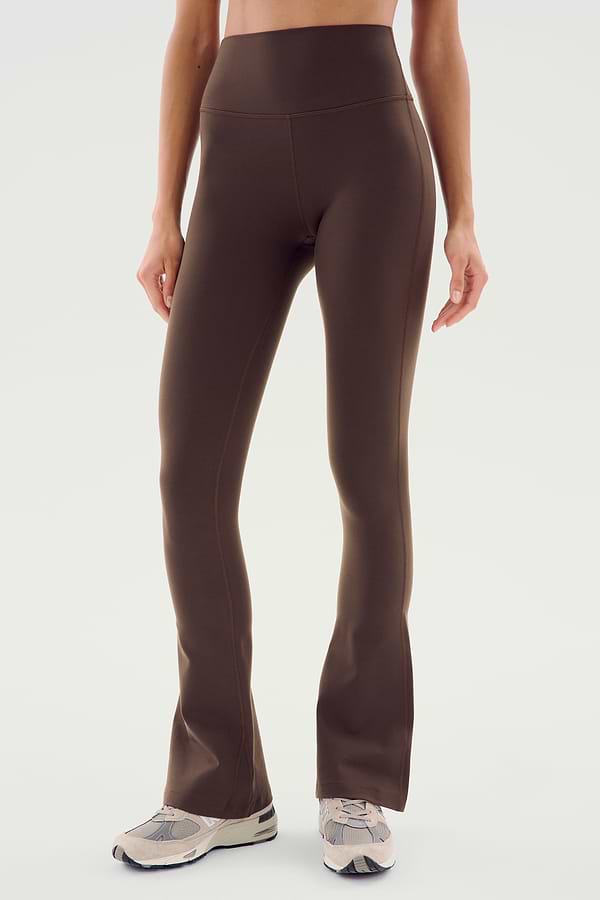 A person wearing SPLITS59 Raquel High Waist Flared Legging in Dark Chocolate paired with white sneakers stands against a plain light background.