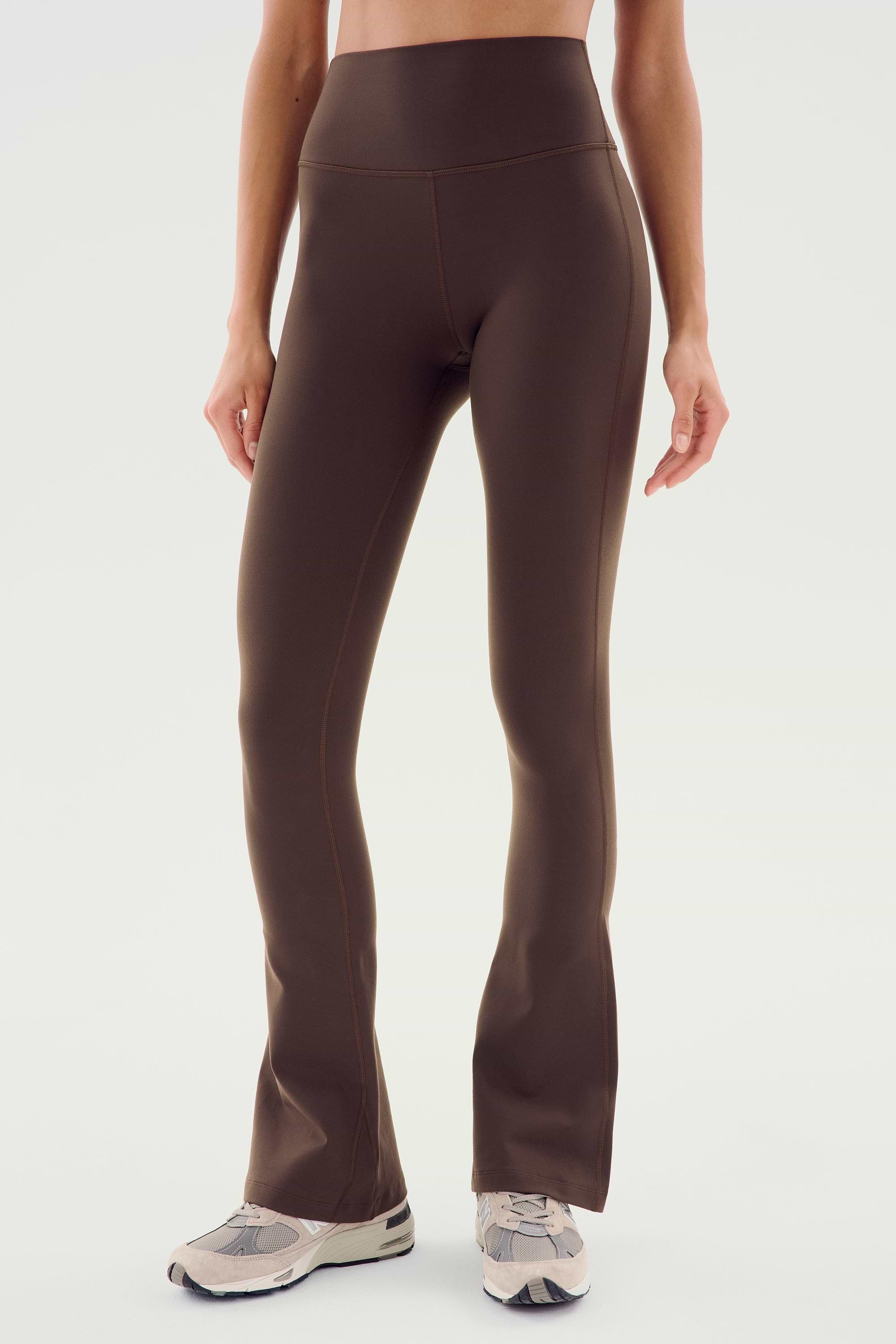A person wearing SPLITS59 Raquel High Waist Flared Legging in Dark Chocolate paired with white sneakers stands against a plain light background.