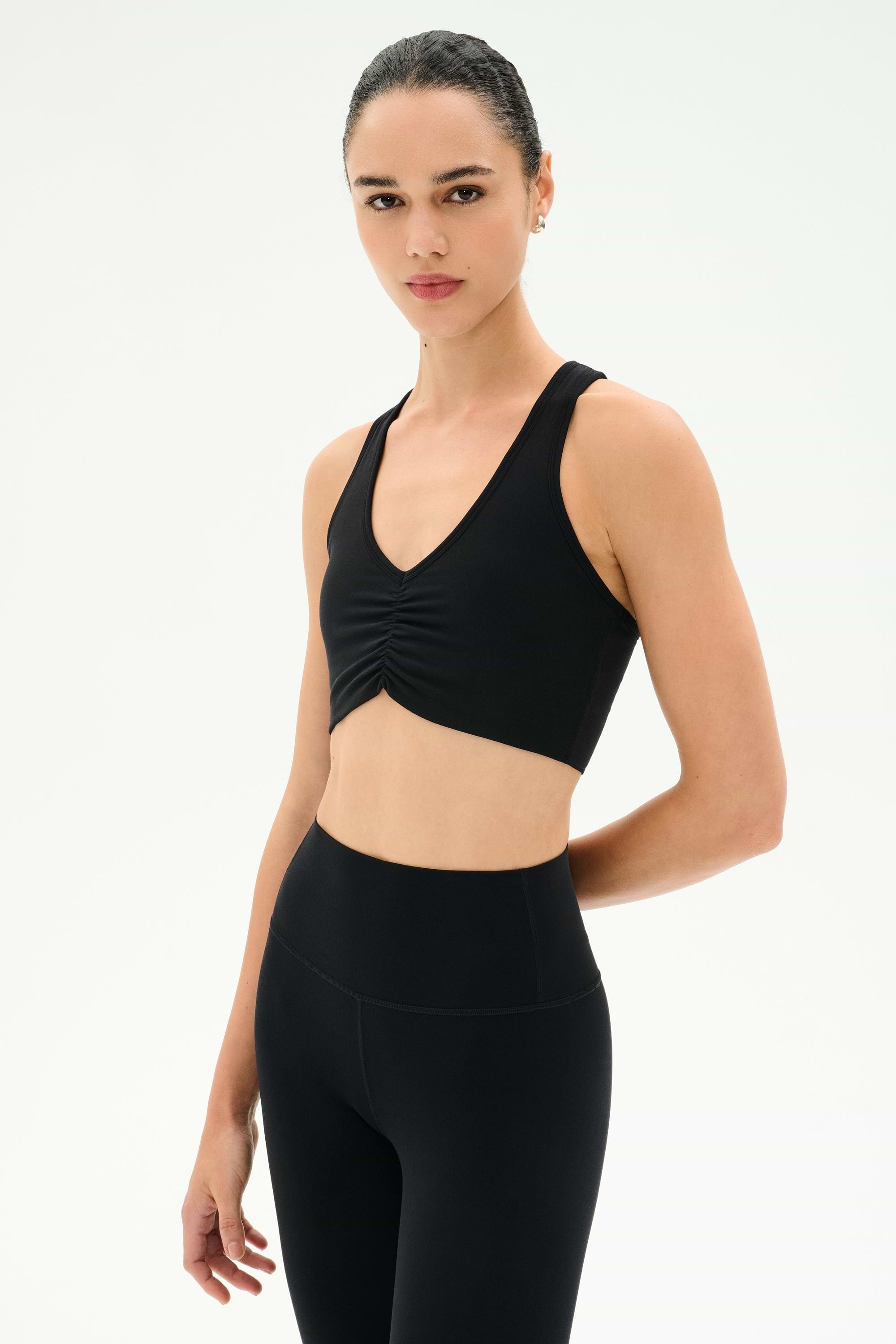 A woman models the SPLITS59 Airweight Bra with Ruching in black, paired with black high-waisted leggings, standing against a white background—perfect attire for hot yoga or any workout.