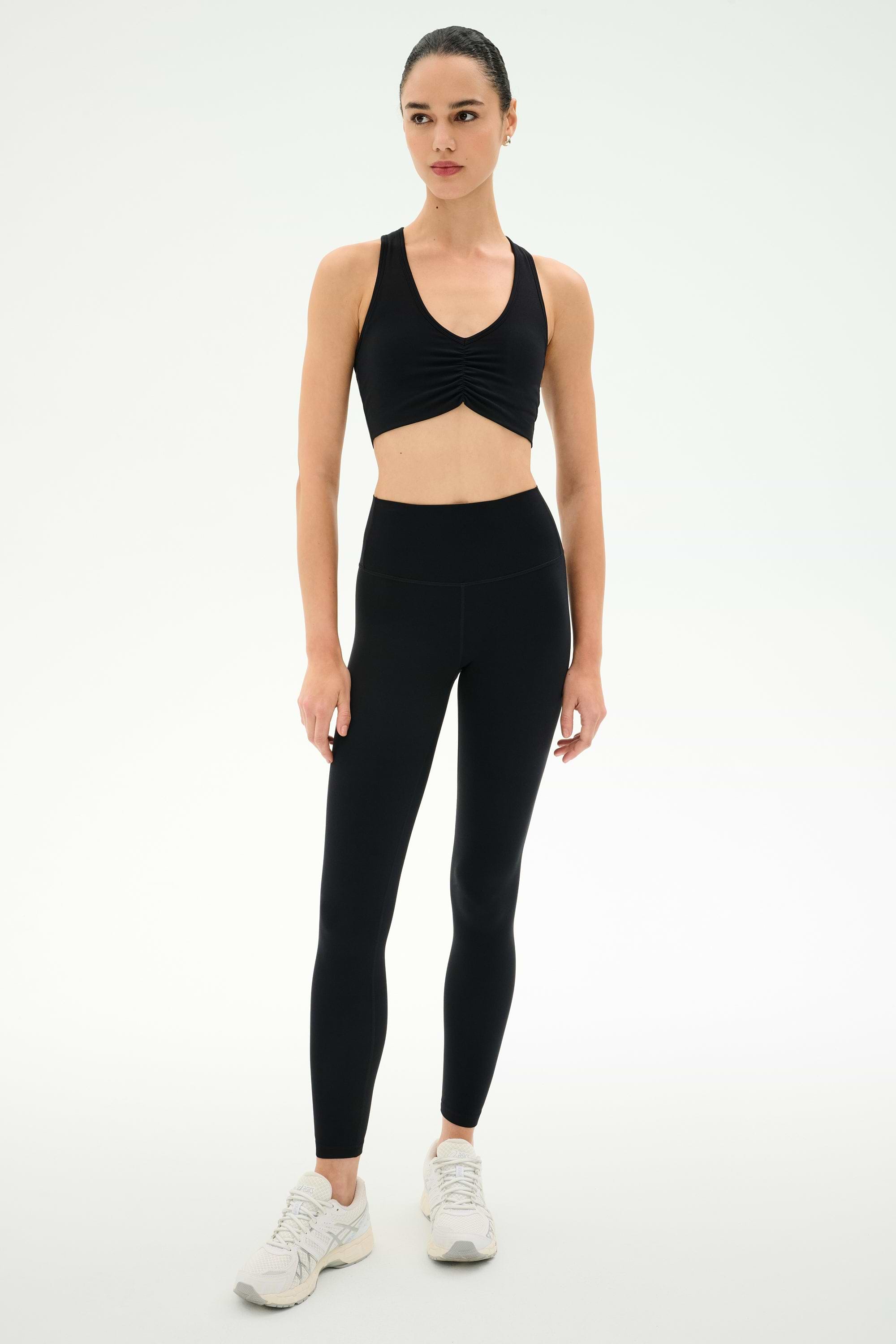 Model wearing SPLITS59 Airweight Bra with Ruching in black, paired with high-waisted black leggings and white athletic shoes, poses against a white background—ideal activewear for hot yoga sessions.