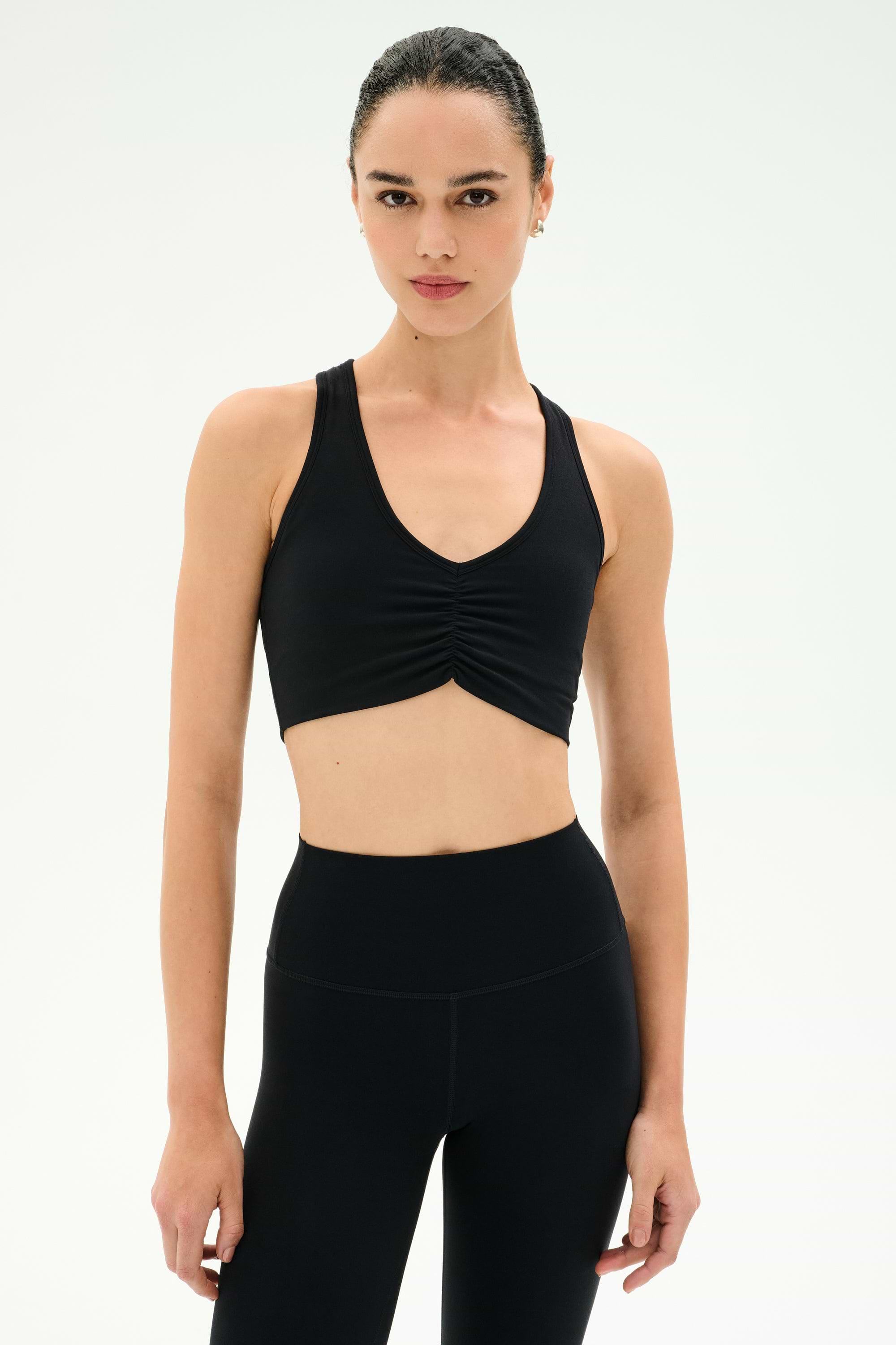 A woman models the SPLITS59 Airweight Bra with Ruching in black, ideal for hot yoga, paired with high-waisted leggings against a plain white background.