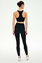 A person stands facing away, wearing the SPLITS59 Airweight Bra with Ruching in black and high-waisted leggings—ideal for hot yoga—with white athletic shoes against a plain light background.