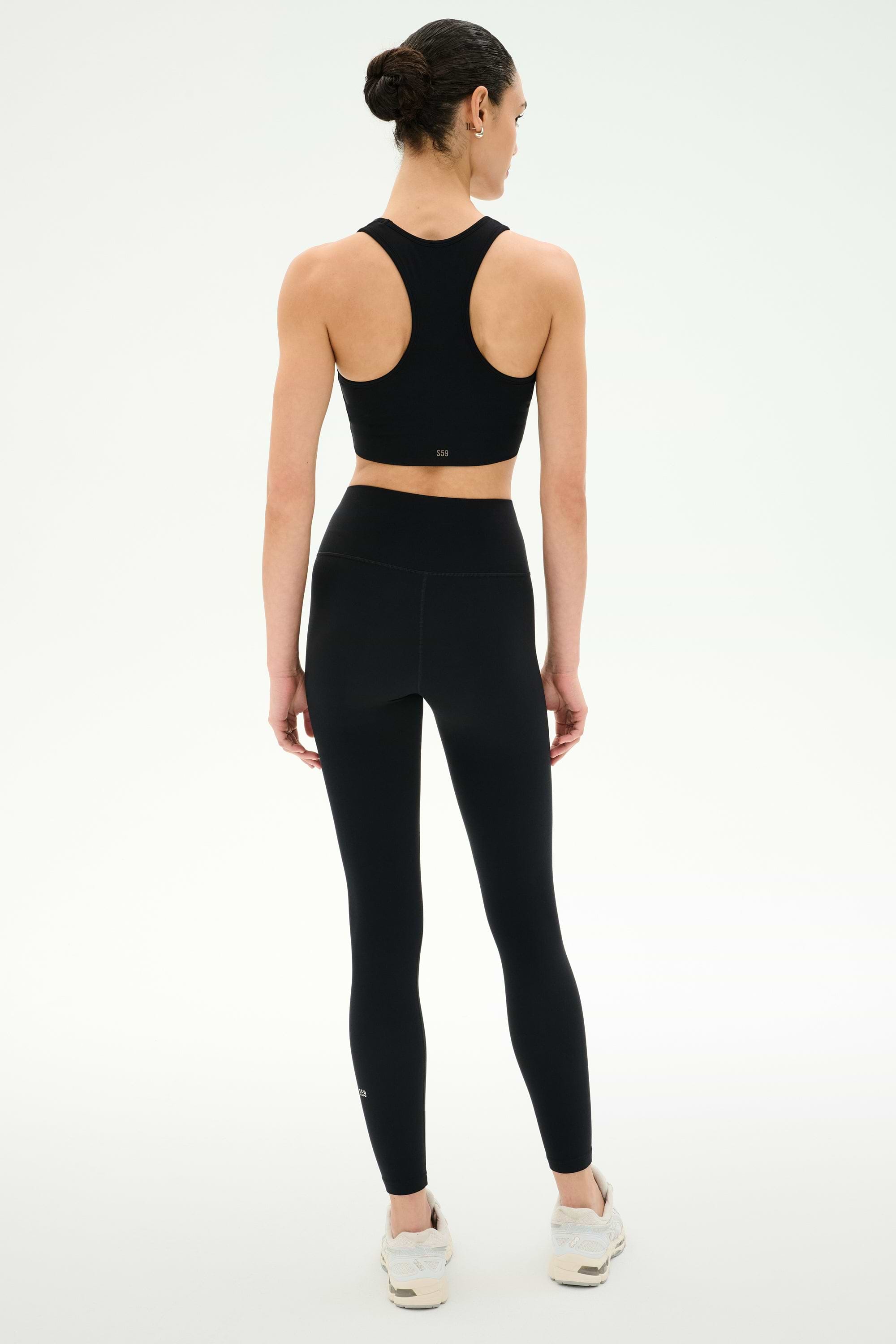 A person stands facing away, wearing the SPLITS59 Airweight Bra with Ruching in black and high-waisted leggings—ideal for hot yoga—with white athletic shoes against a plain light background.