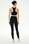 A person stands facing away, wearing the SPLITS59 Airweight Bra with Ruching in black and high-waisted leggings—ideal for hot yoga—with white athletic shoes against a plain light background.