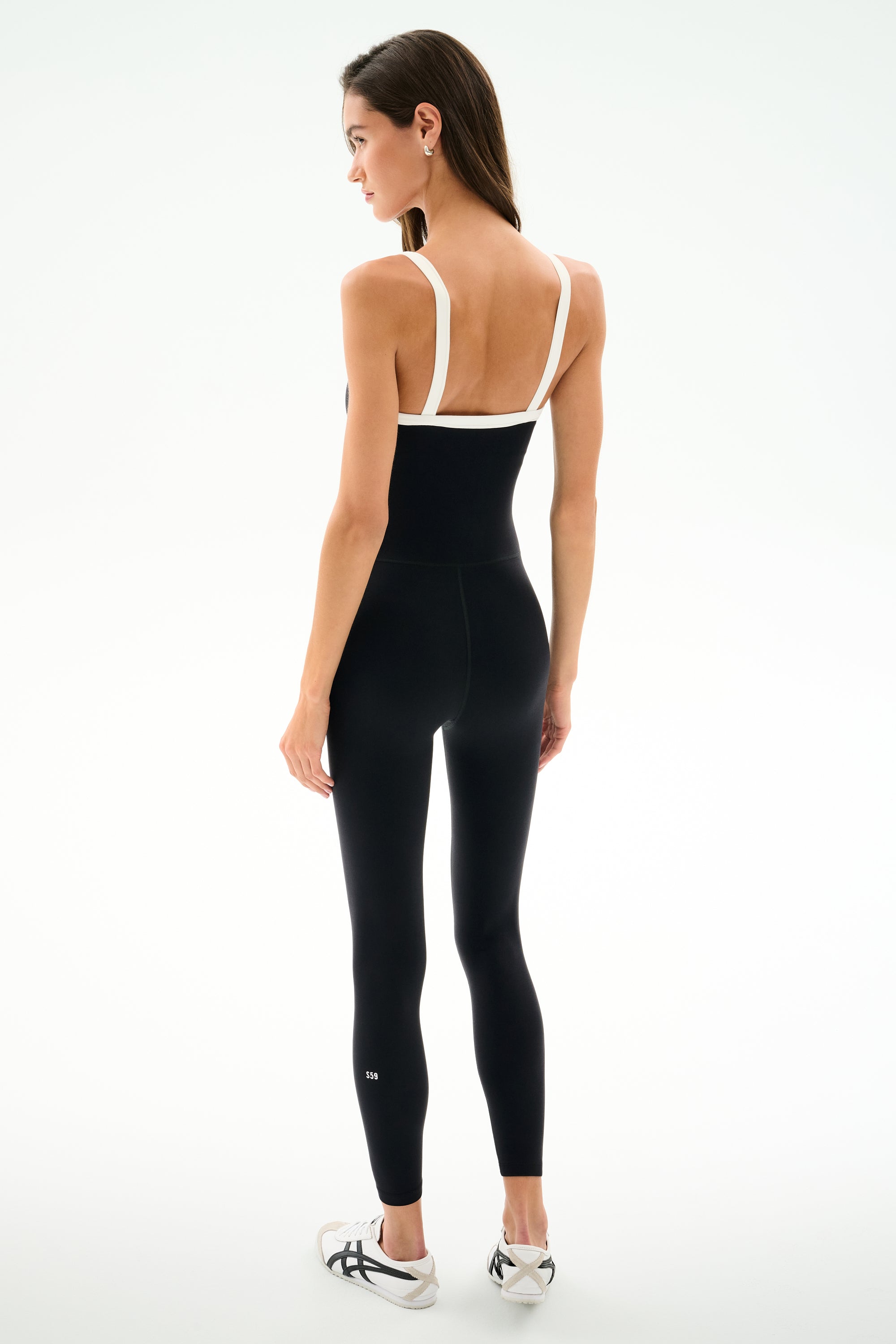 A woman stands facing away in the SPLITS59 Monah Rigor Jumpsuit 7/8 - Black/White with white sneakers, set against a plain white background.
