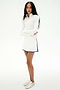 A woman stands with arms crossed, wearing the SPLITS59 Ozzie Rigor Skort in White/Cobalt, a white long-sleeve zip-up top, and white sneakers against a plain white background.