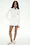 A woman stands against a white background, wearing the SPLITS59 Ozzie Rigor Skort in White/Cobalt with built-in shorts, paired with a long-sleeve white athletic top and white sneakers—ideal for court sports.