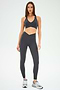 A woman stands against a plain background, dressed in SPLITS59’s Mia Rigor 7/8 Leggings in Graphite, crafted from durable Rigor fabric and paired with a matching dark gray sports bra and gray sneakers.
