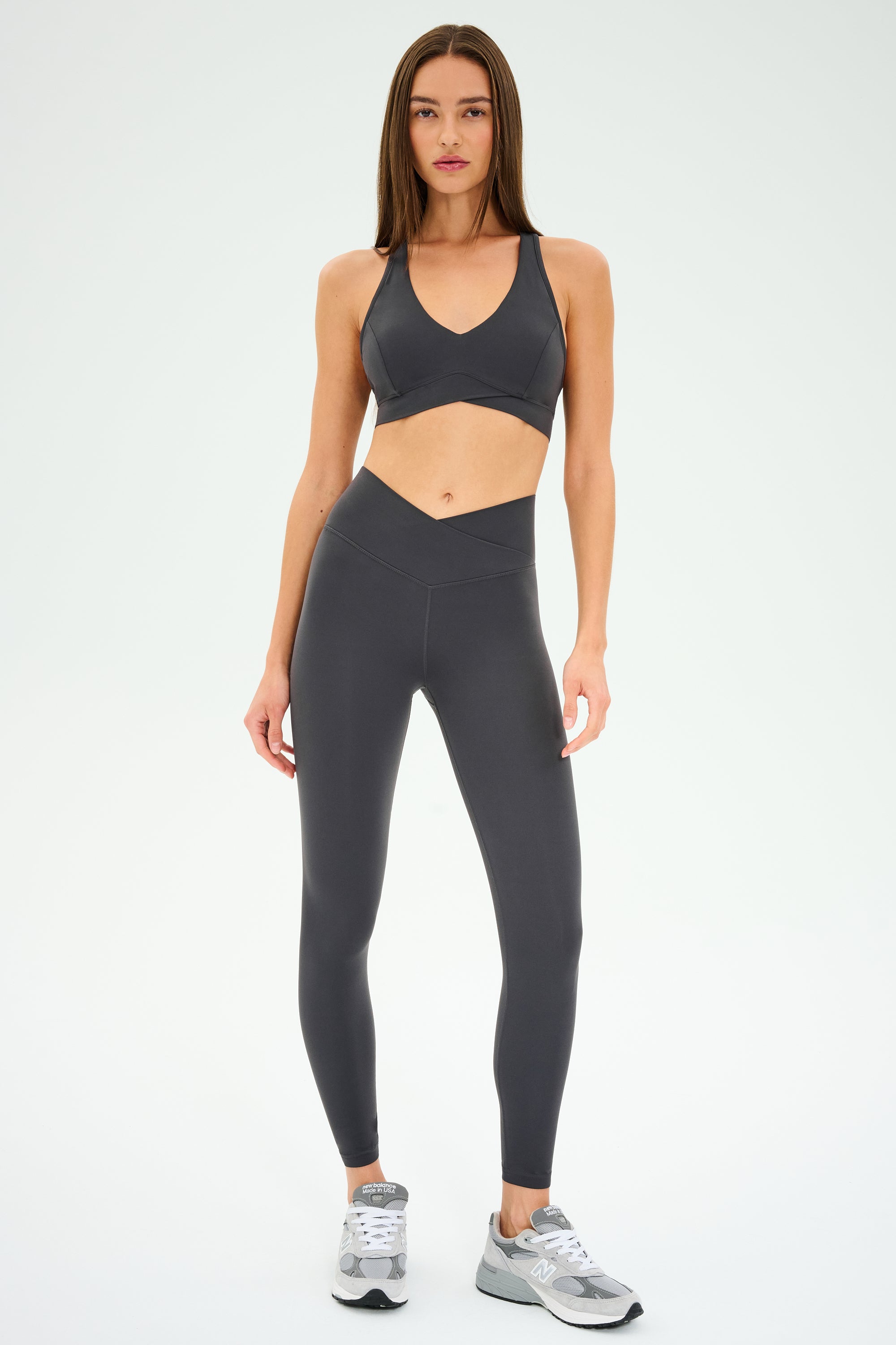 A woman stands on a white background wearing the SPLITS59 Mia Rigor Bra in Graphite, paired with matching high-waisted leggings and gray sneakers—an outfit designed for multi-sport performance and ideal for gym workouts.