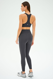 A woman, viewed from the back against a white background, wears the SPLITS59 Mia Rigor 7/8 - Graphite high waist leggings crafted from Rigor fabric for superior support, paired with a black sports bra and gray athletic shoes.