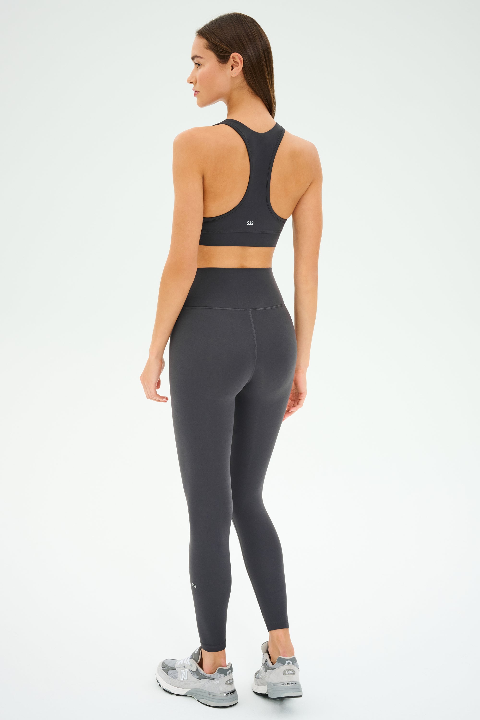 A woman stands on a white background, facing away, wearing the SPLITS59 Mia Rigor Bra in Graphite—a black racerback bra designed for multi-sport performance—paired with matching high-waisted leggings and gray sneakers, making it perfect for gym workouts.