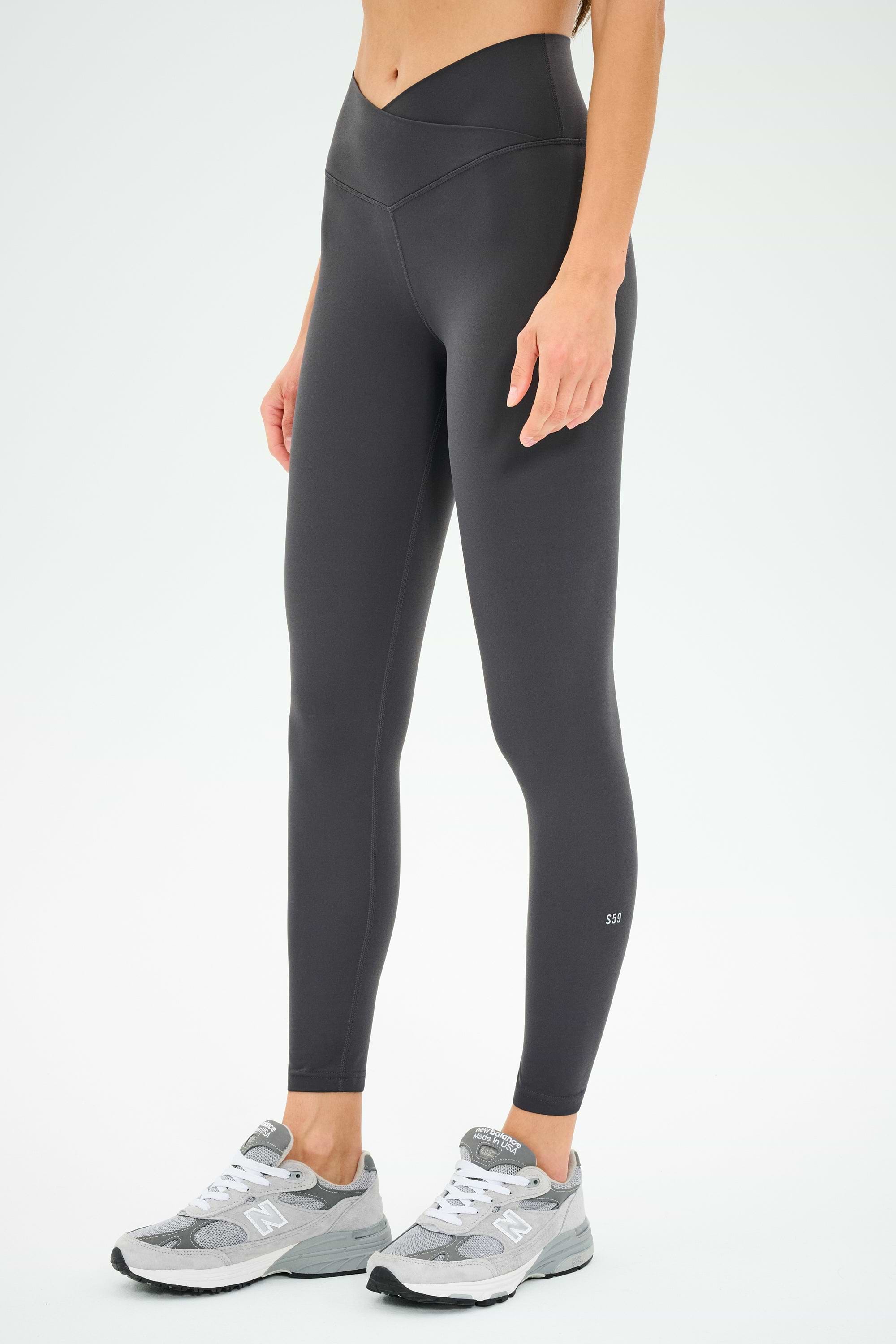 A person wearing SPLITS59 Mia Rigor 7/8 leggings in Graphite, paired with gray sneakers, stands against a plain white background.