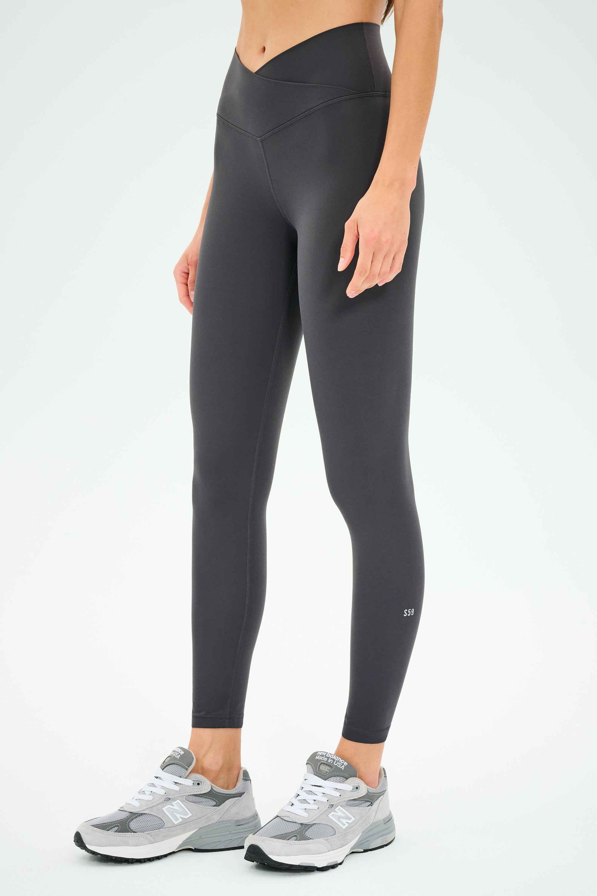A person wearing SPLITS59 Mia Rigor 7/8 leggings in Graphite, paired with gray sneakers, stands against a plain white background.