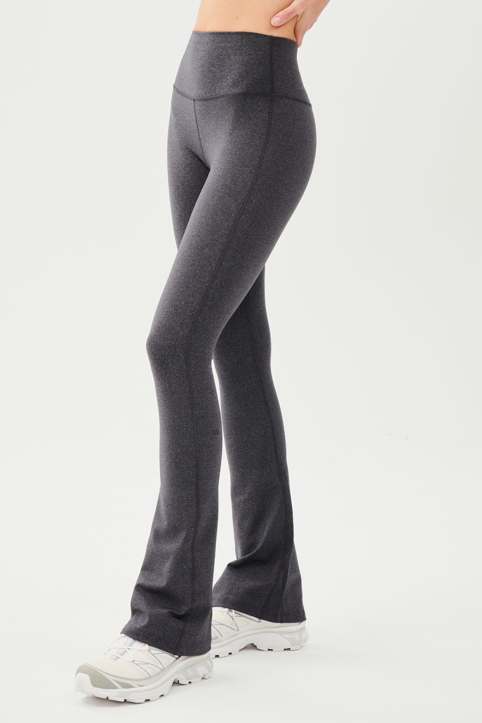 Front side view of woman wearing dark gray high waist below ankle length legging with wide flared bottoms. Paired with white shoes.