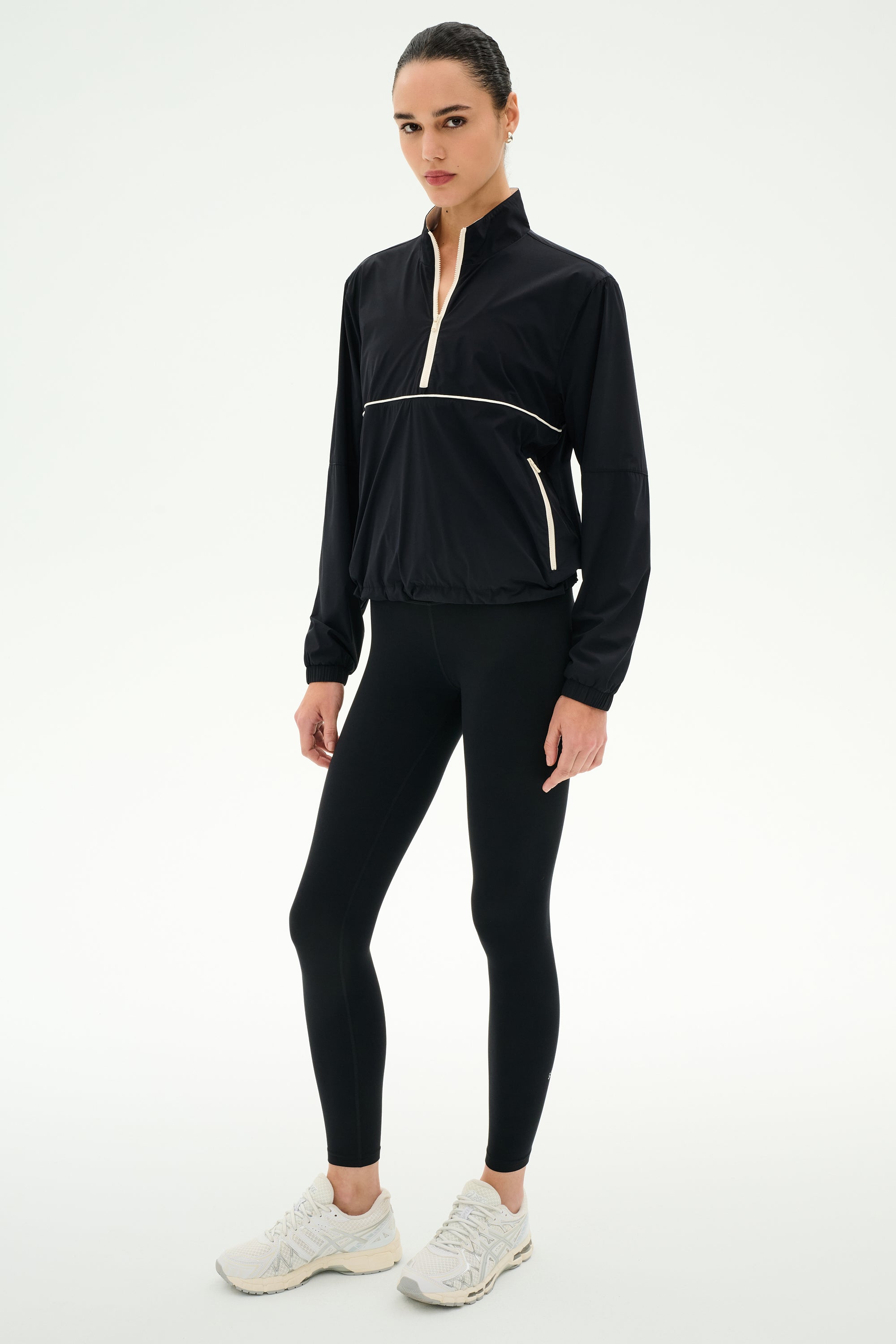 A woman wears the SPLITS59 Lyle Woven Nylon Pullover in Black/Creme, styled with spandex leggings and white sneakers, standing against a plain white background.