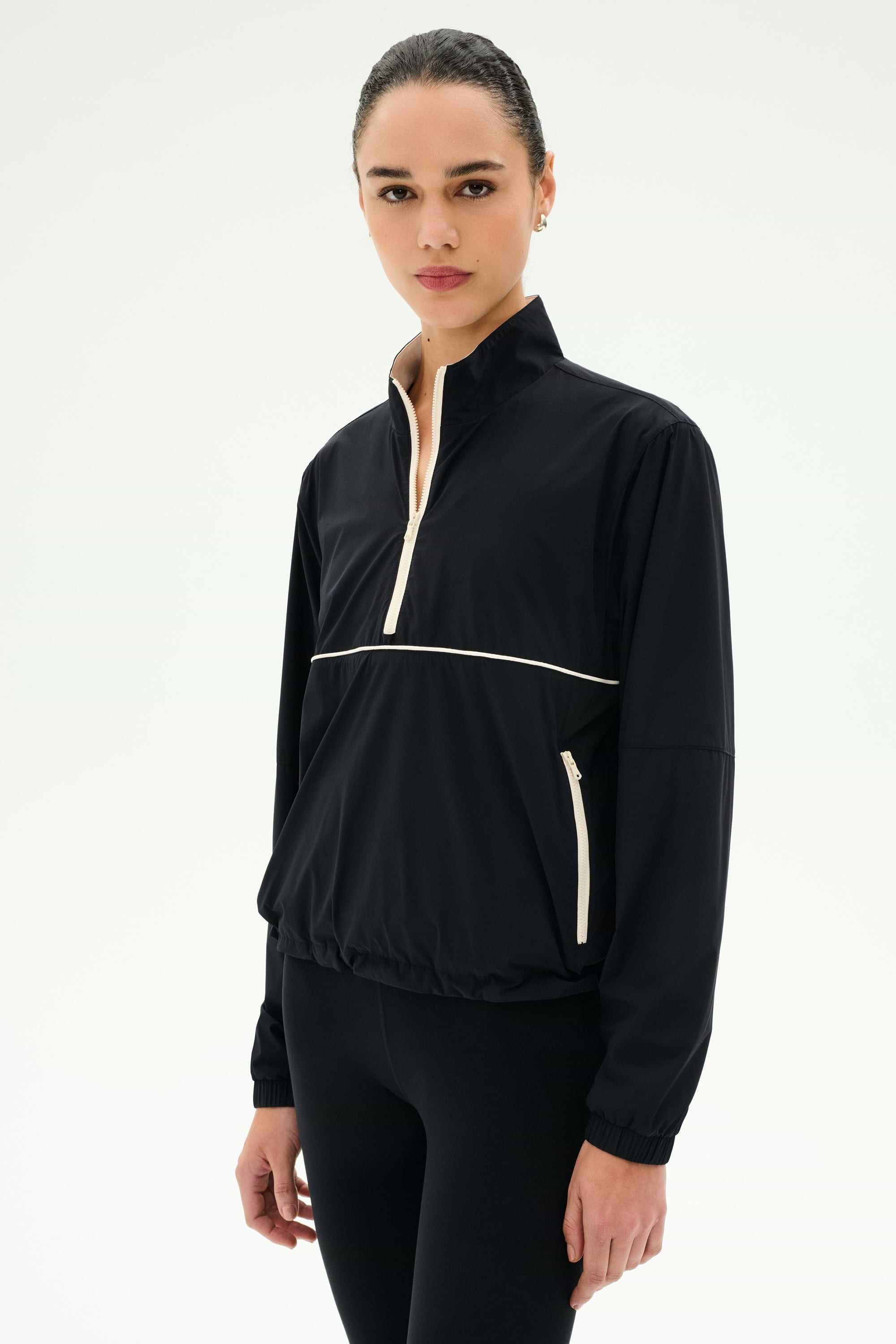 A woman wears the SPLITS59 Lyle Woven Nylon Pullover in Black/Creme with matching black spandex pants, standing against a plain white background.