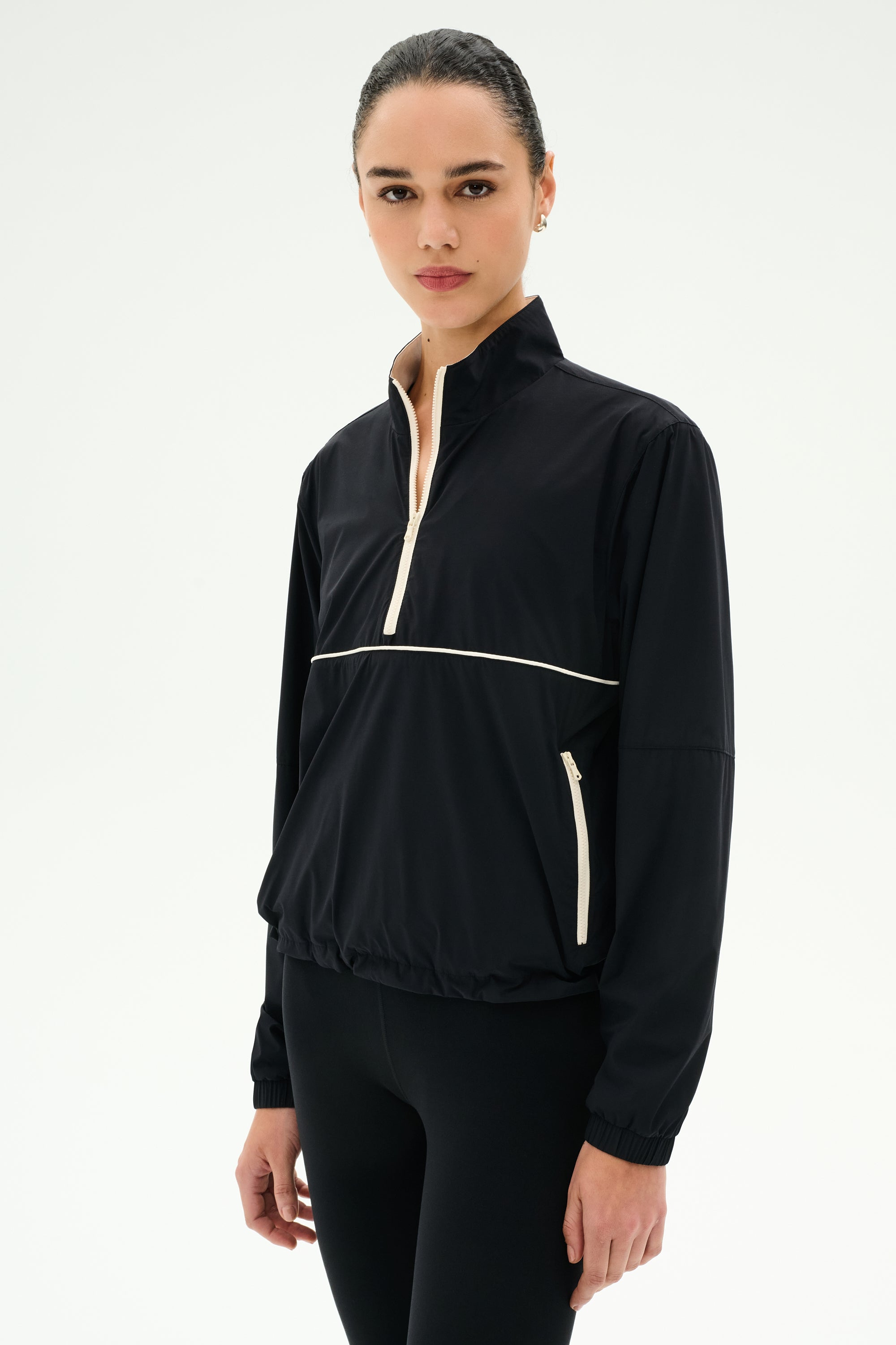 A woman wears the SPLITS59 Lyle Woven Nylon Pullover in Black/Creme with matching black spandex pants, standing against a plain white background.