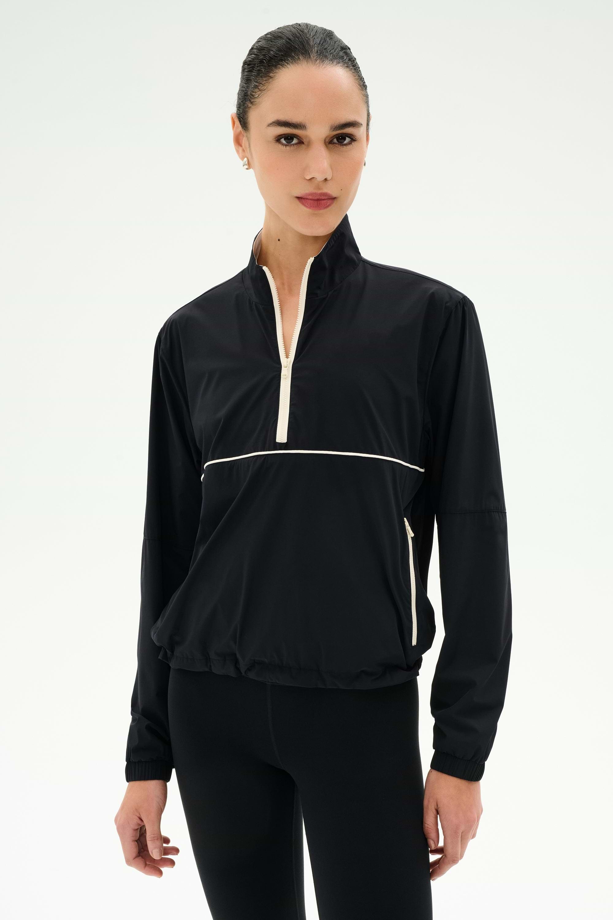 A woman wears the SPLITS59 Lyle Woven Nylon Pullover in Black/Creme with black spandex leggings, standing against a plain white background.