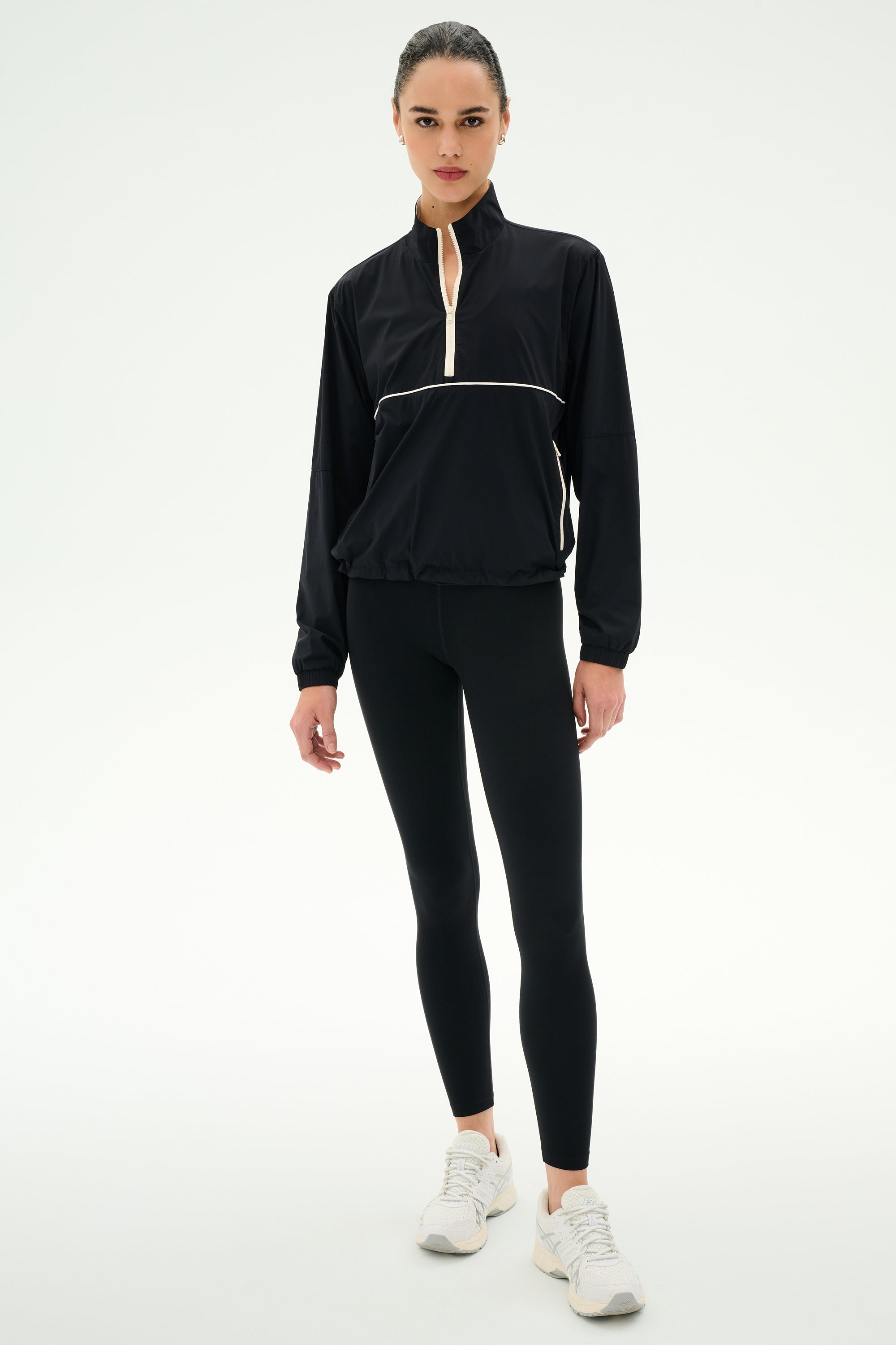 A person wearing the SPLITS59 Lyle Woven Nylon Pullover in Black/Creme, black spandex leggings, and white sneakers stands against a plain white background.